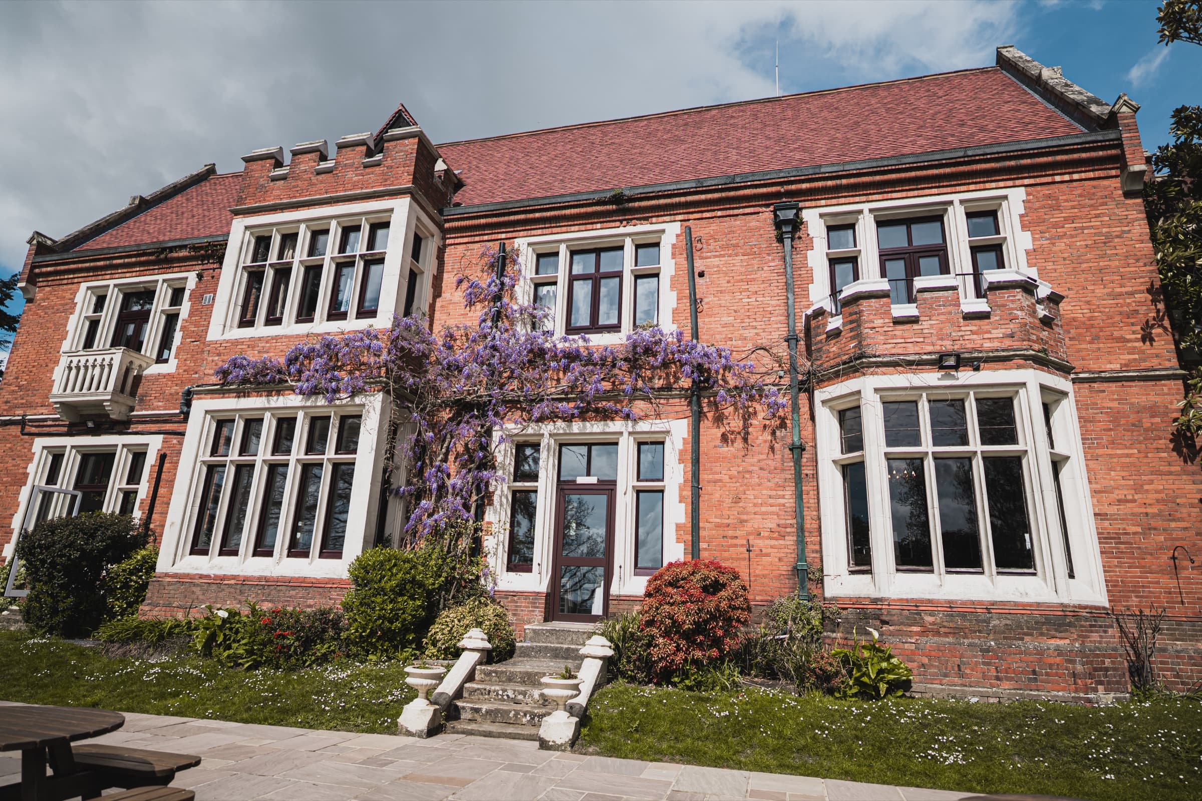Red-brick Victorian facade of Highley Manor clad in flowering purple wisteria on a wedding day