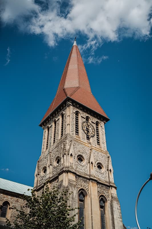 The flint-and-stone clock tower and red-tiled spire of St John’s Church, East Dulwich, against a blue sky