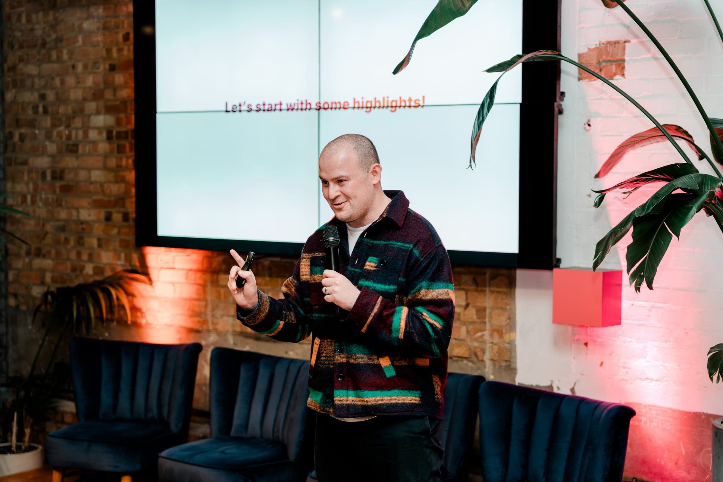 A speaker mid-talk with a microphone at a relaxed corporate event in an exposed-brick London venue