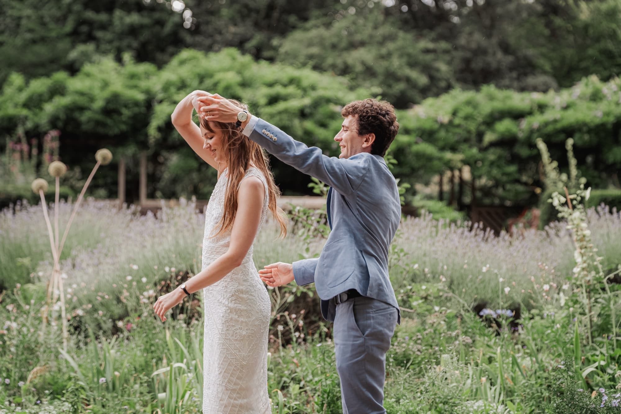 Bride in a white lace dress spins beneath the groom’s raised hand in a tall lavender and wildflower meadow, both laughing