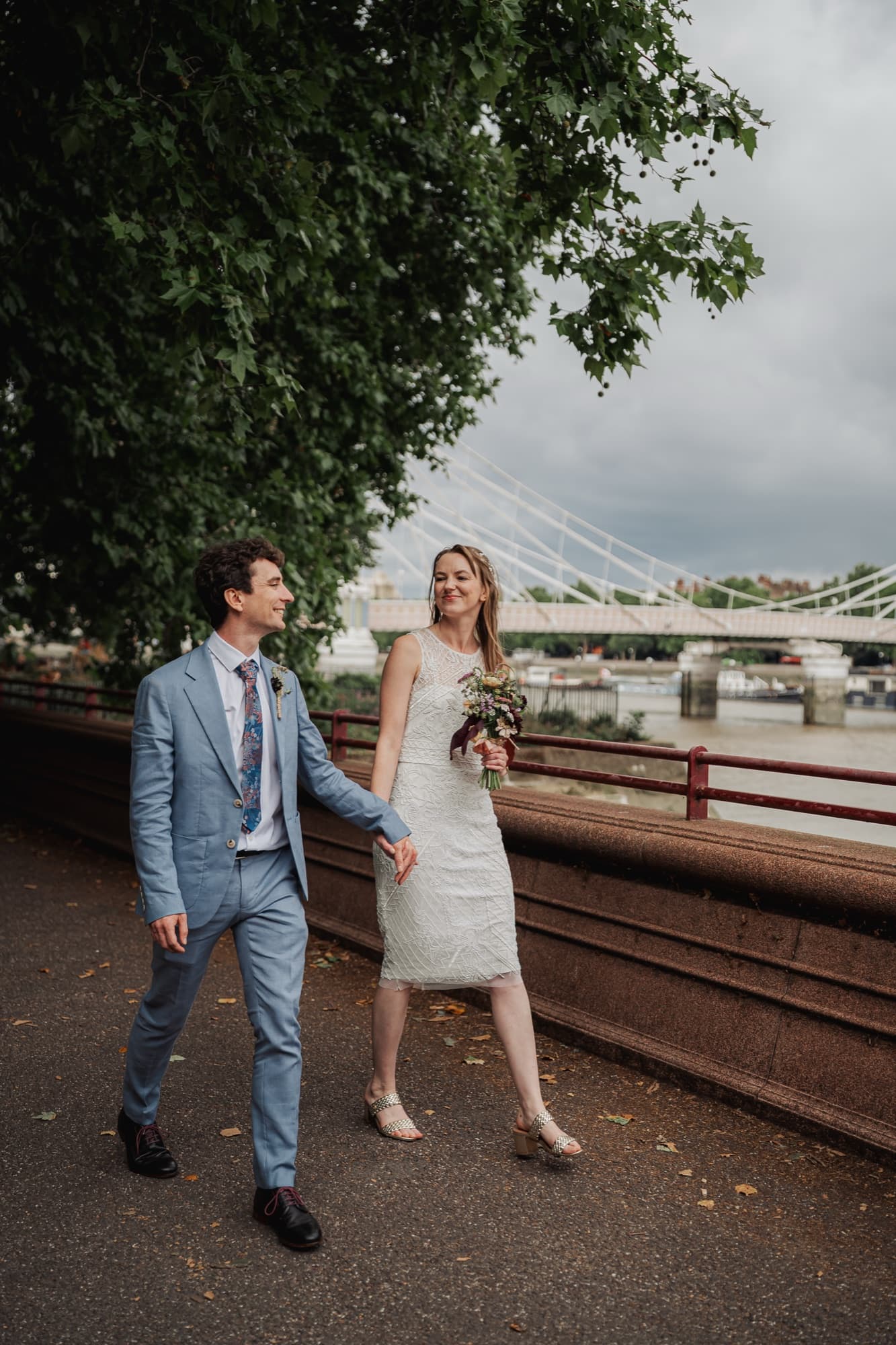 Bride and groom walk hand in hand along the Thames path with the towers of Albert Bridge behind them