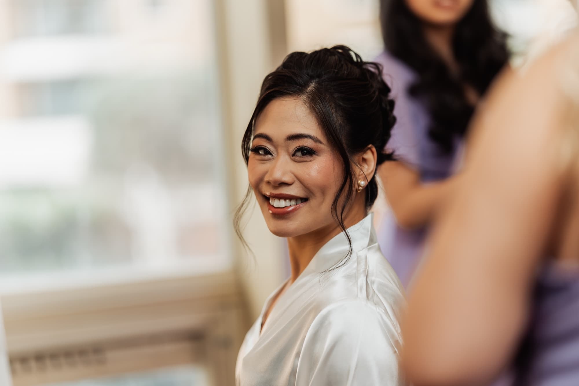 A bride in a white satin robe glances over her shoulder with a radiant smile in soft window light, bridesmaids behind