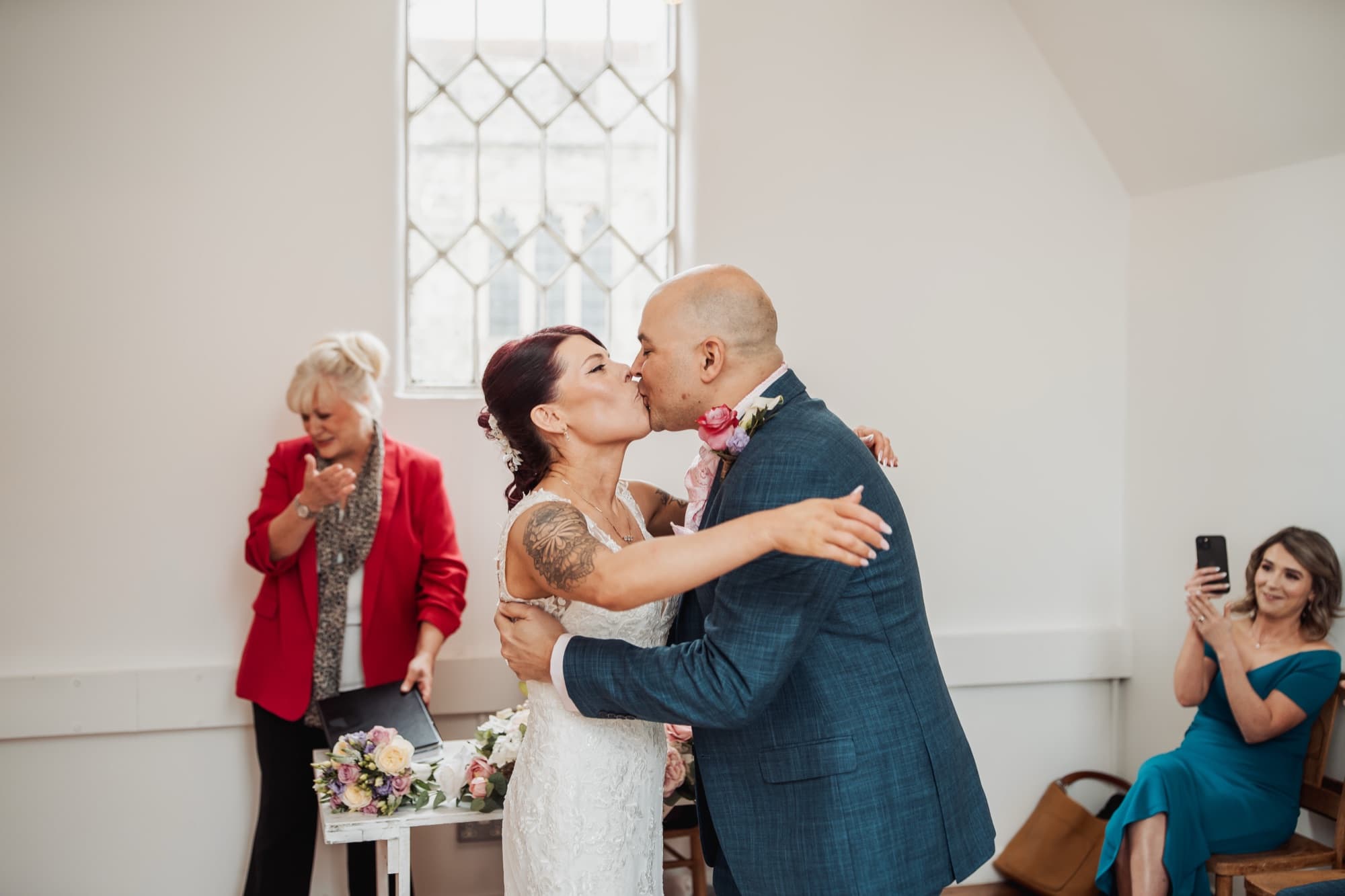 A couple share their first kiss in a bright white ceremony room as guests clap, soft daylight from a tall arched window behind