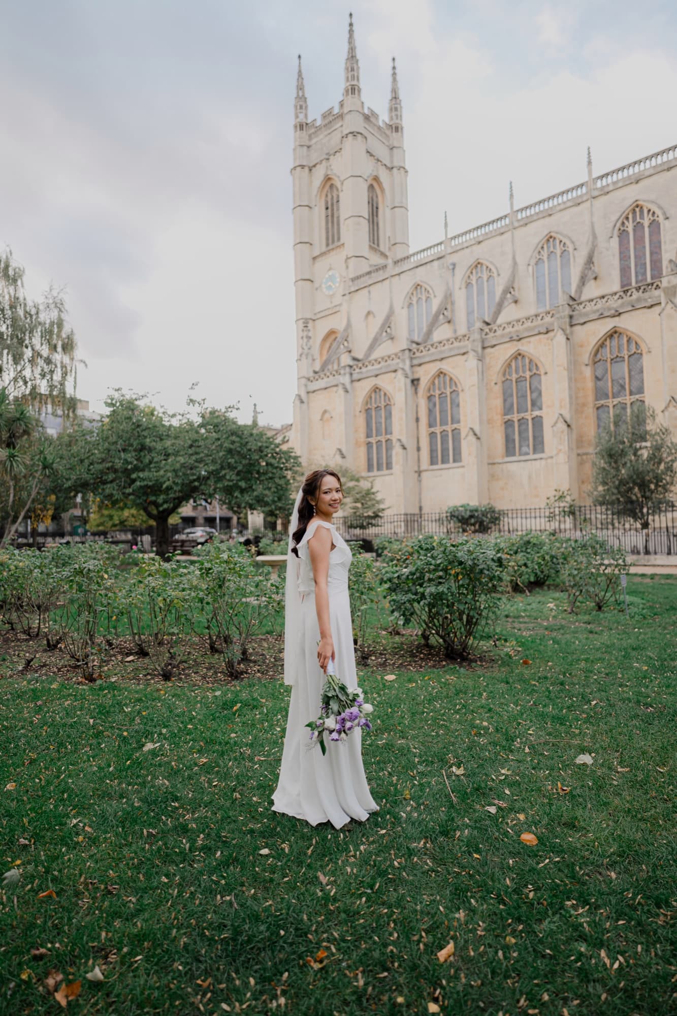 Bride glancing back over her shoulder in a flowing gown, bouquet at her side, a Gothic chapel framed behind her