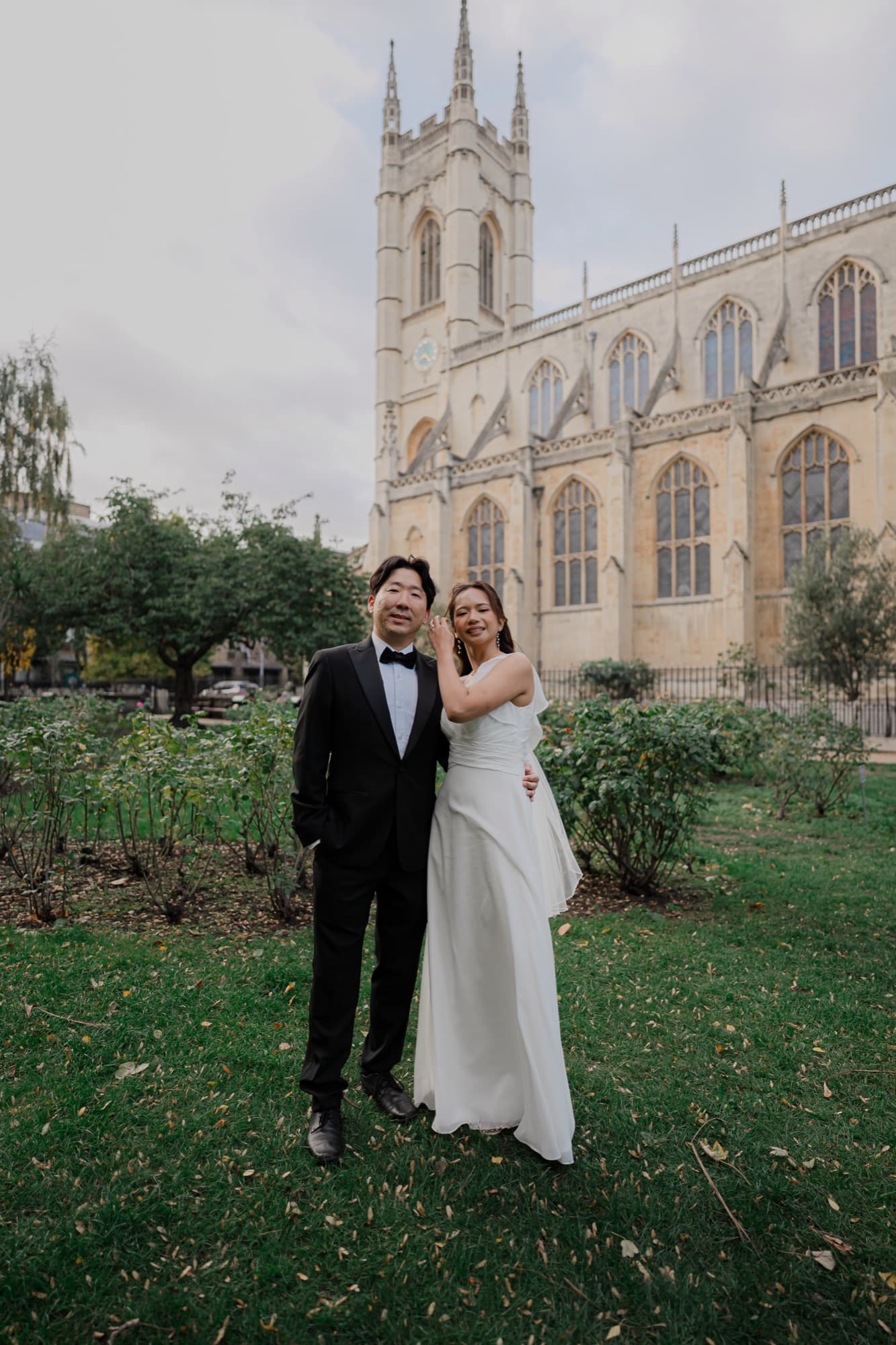 Bride and groom standing close in a garden with a soaring Gothic stone chapel and tower behind them