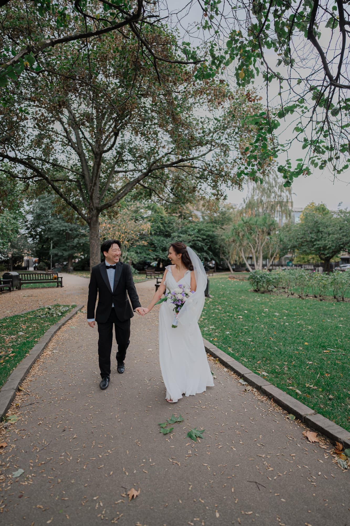Bride and groom holding hands as they walk a tree-lined garden path, the bride laughing in a flowing gown and veil