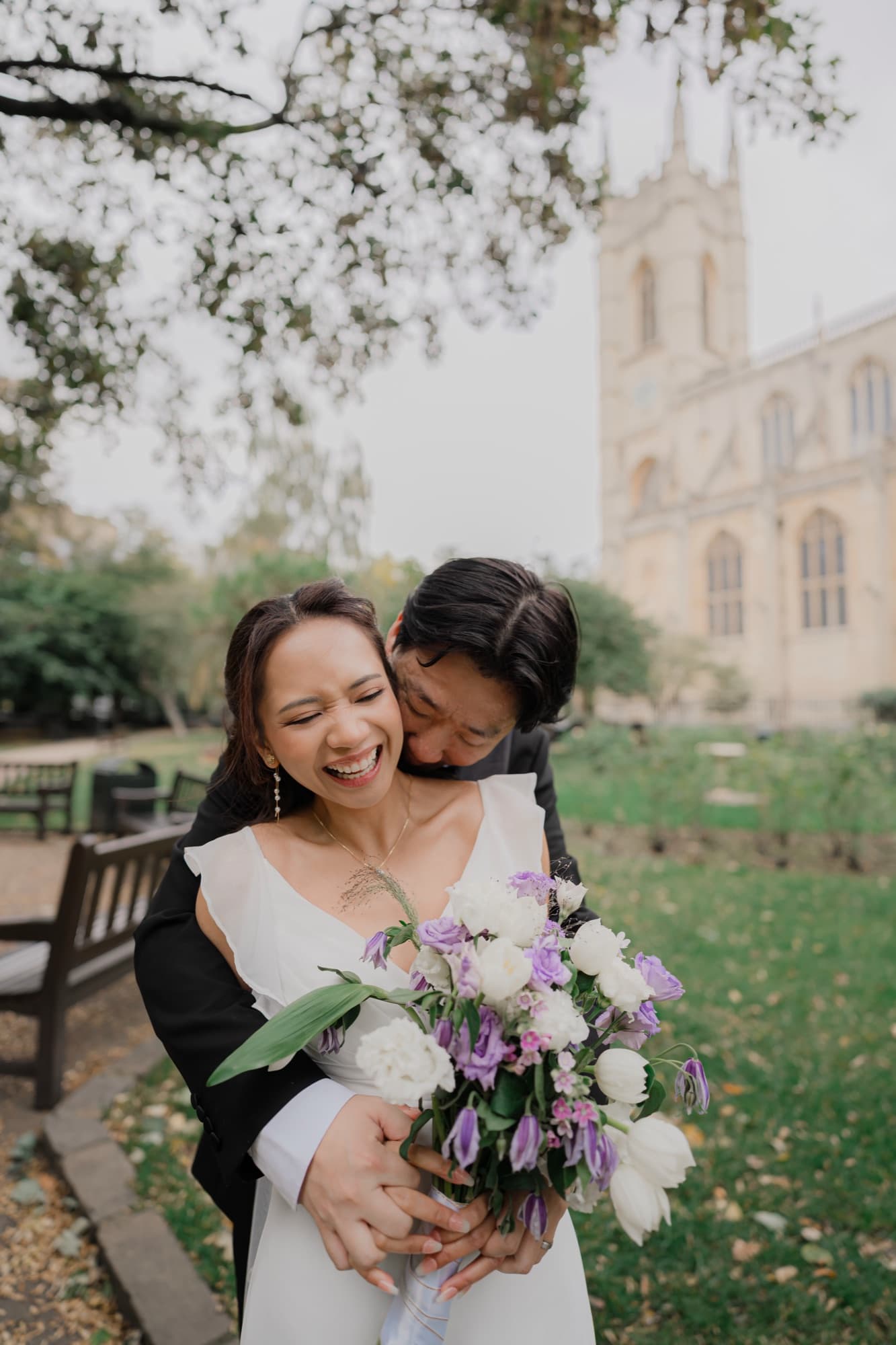 Groom wrapping his arms around his laughing bride from behind as she holds a white and lilac bouquet, a Gothic chapel rising softly behind