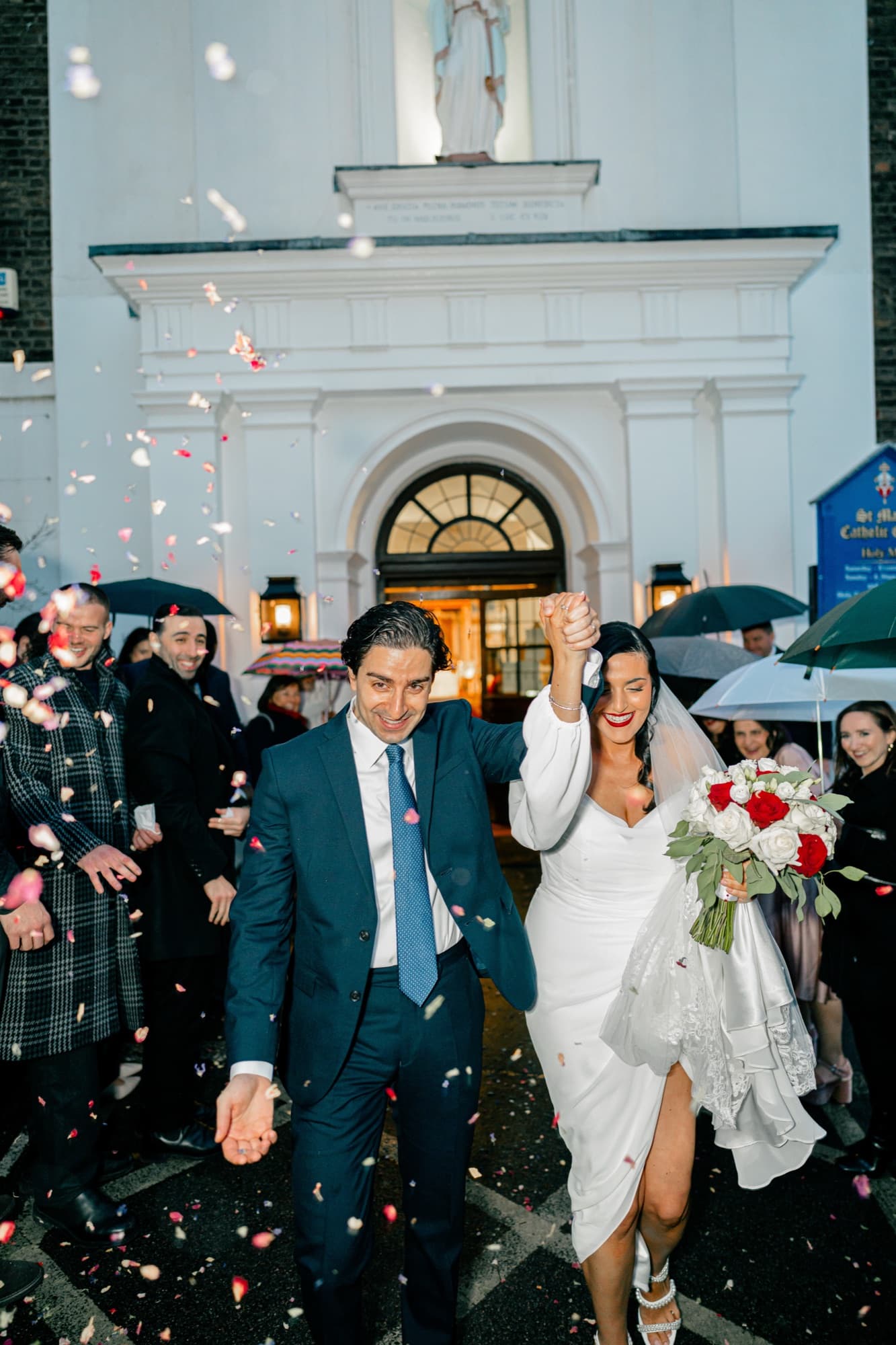 Newlyweds raise their joined hands and stride through a shower of confetti outside a white church, guests cheering with umbrellas