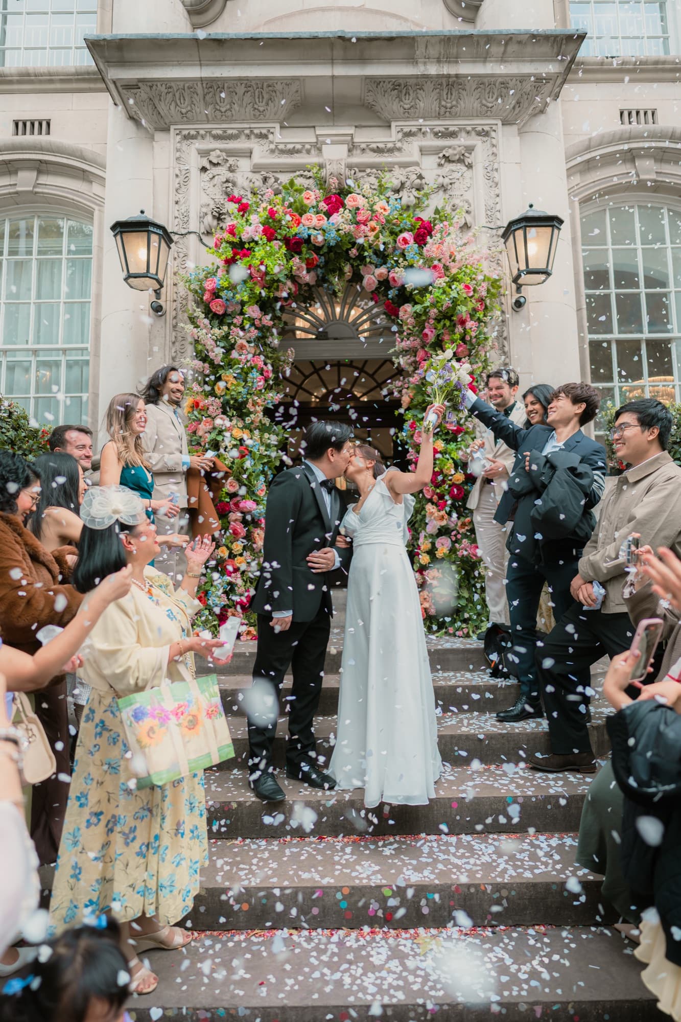 A couple kiss on the venue steps beneath a lavish floral arch as guests throw confetti outside a grand London townhouse