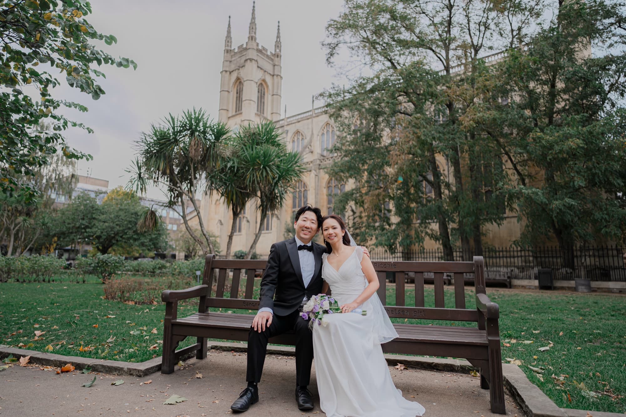A couple seated together on a park bench with a Gothic stone chapel rising behind them in soft daylight