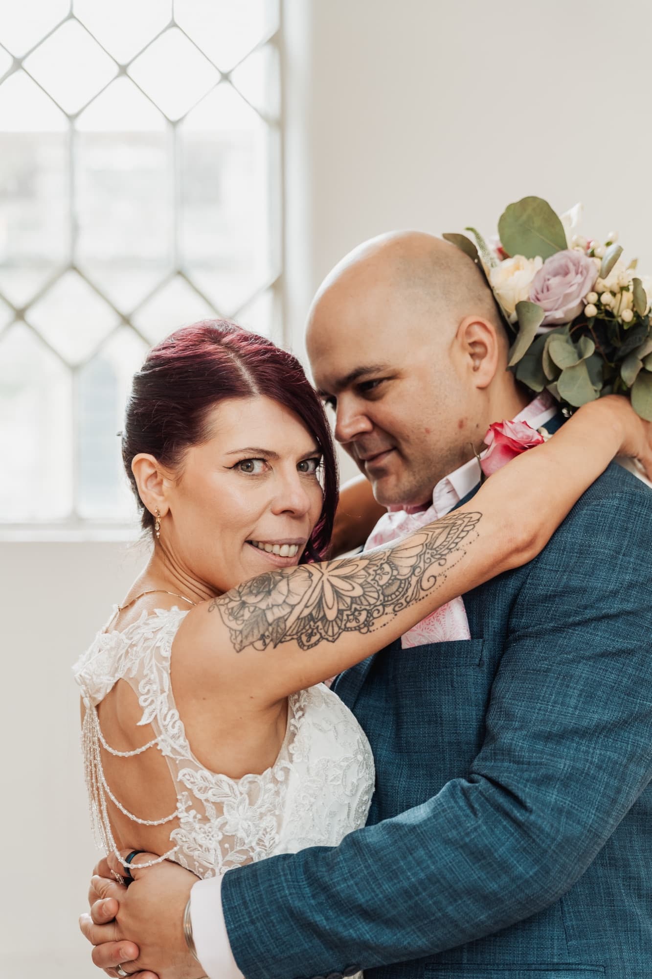 Newlyweds in a close embrace beside a leaded window, the bouquet lifted over a shoulder in soft natural light