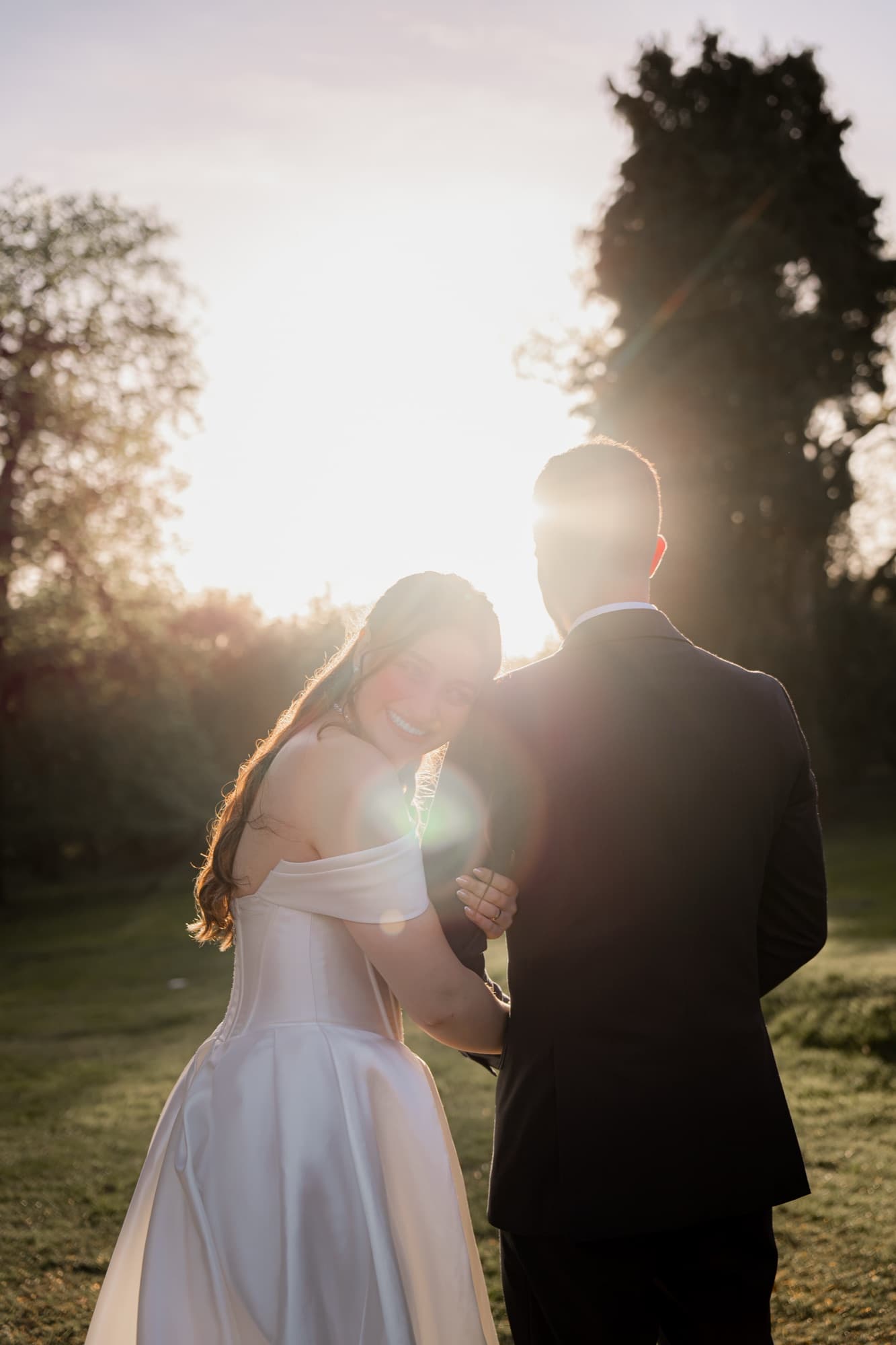 Bride glances back over her shoulder with a warm smile, the sun flaring between her and the groom on the grounds at golden hour