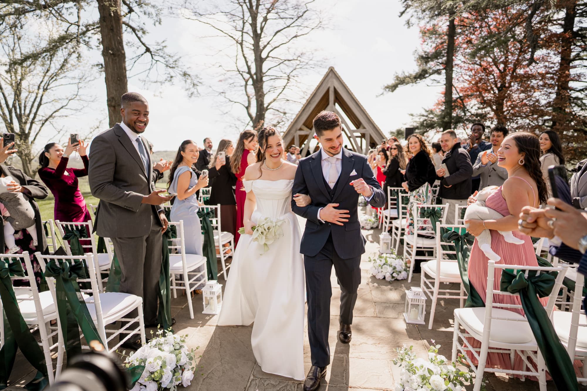 Newlyweds walk arm in arm up the aisle as cheering guests line the chairs, the oak A-frame pavilion behind in spring light