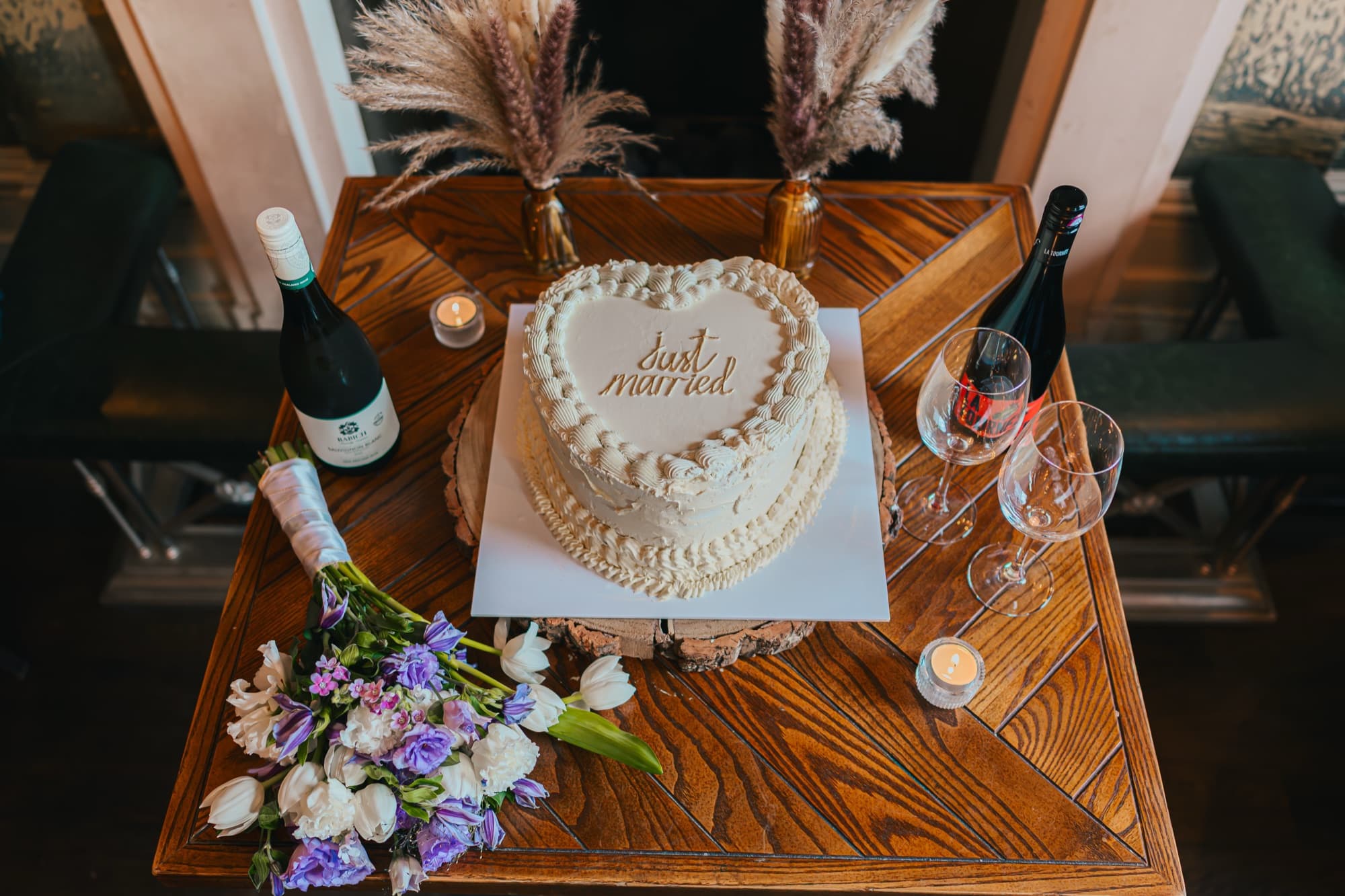 A heart-shaped “just married” cake styled with the bridal bouquet, champagne, candles and dried pampas on a wood table