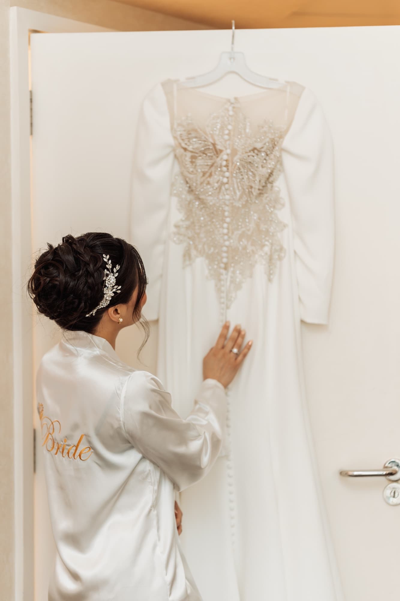A bride reaches up to touch her beaded wedding gown hanging on a door, the long button back in soft light