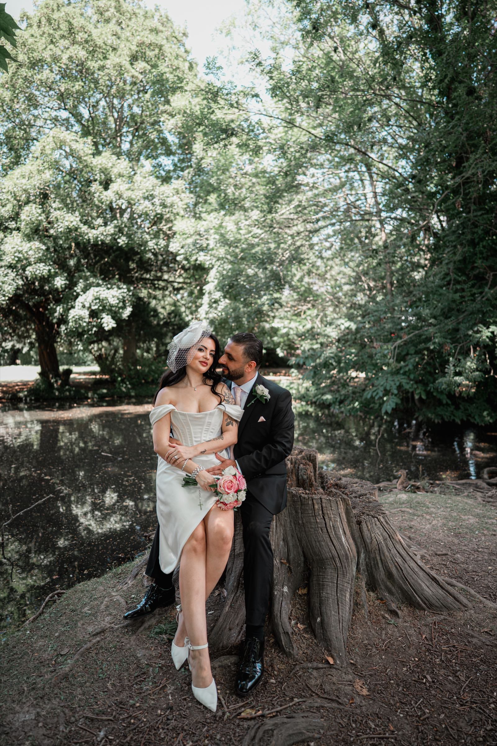 A couple sitting together on a tree stump beside a woodland pond after their wedding