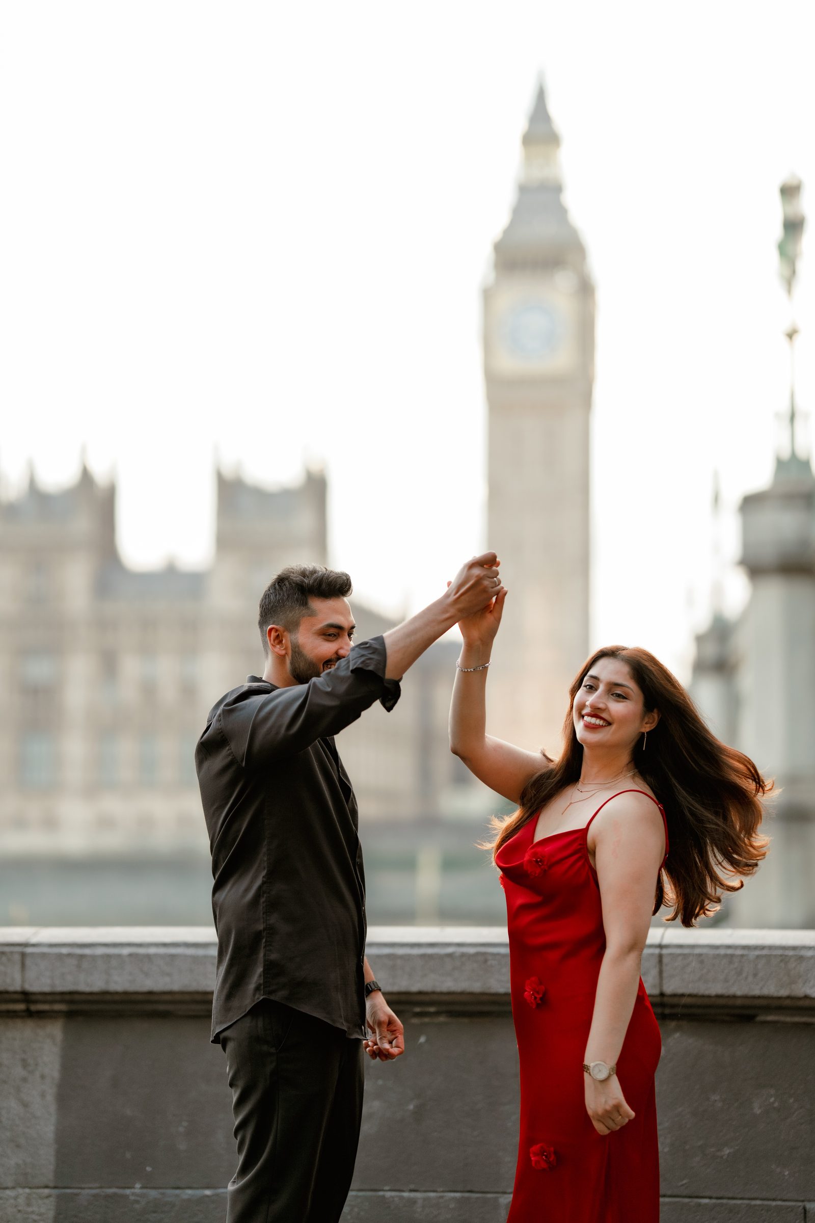 A man twirling his partner in a red dress on the river terrace opposite Big Ben during a London engagement photoshoot
