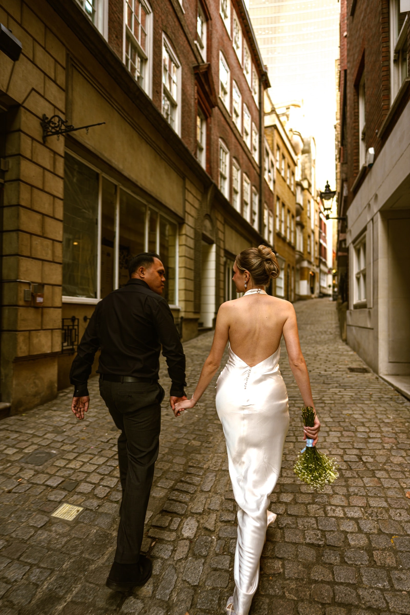 A couple walking hand in hand down a cobbled City of London lane between old brick buildings
