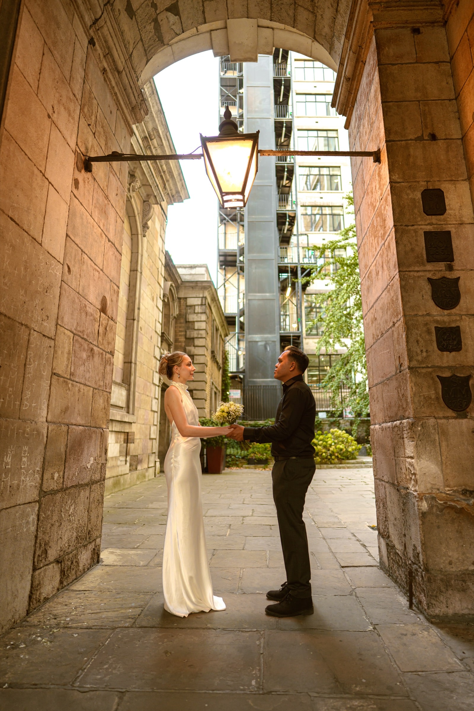 A couple holding hands beneath an old lantern in a stone archway off a City of London lane