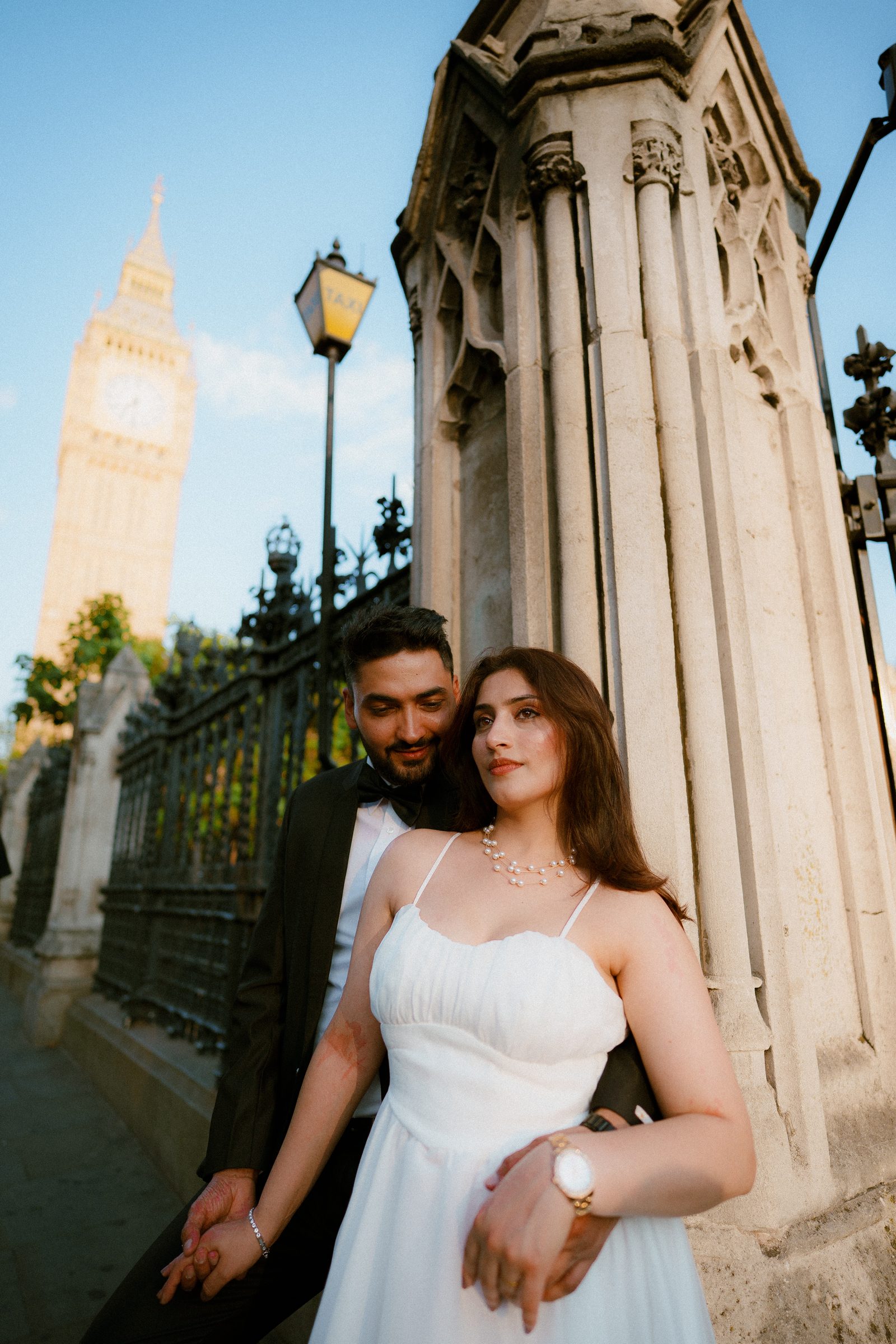 A couple leaning together against the stone gate piers of the Houses of Parliament, a lamp and Big Ben above them