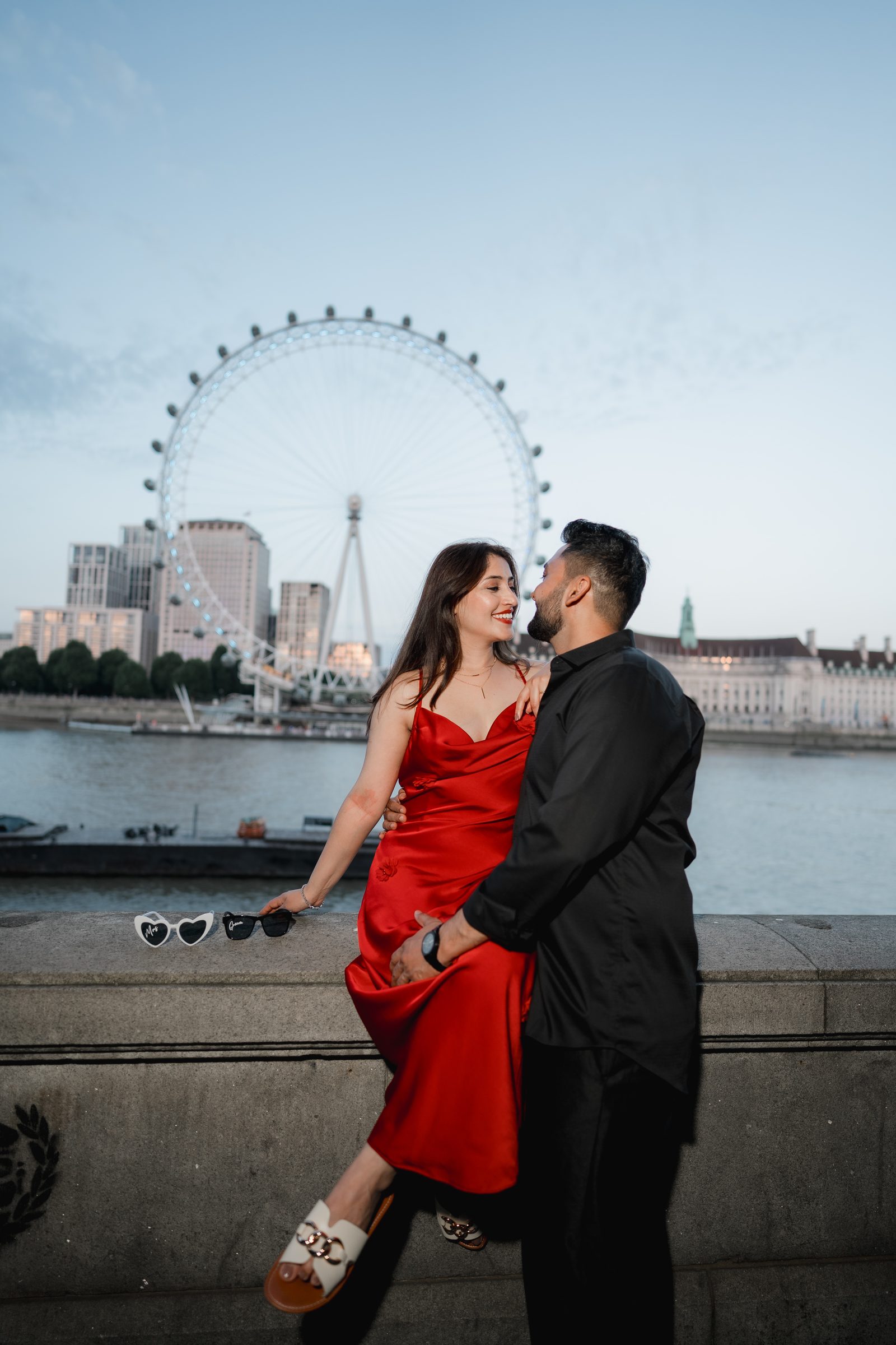 Prachi in heart-shaped sunglasses sitting on the river wall with Rohit, the London Eye rising behind them
