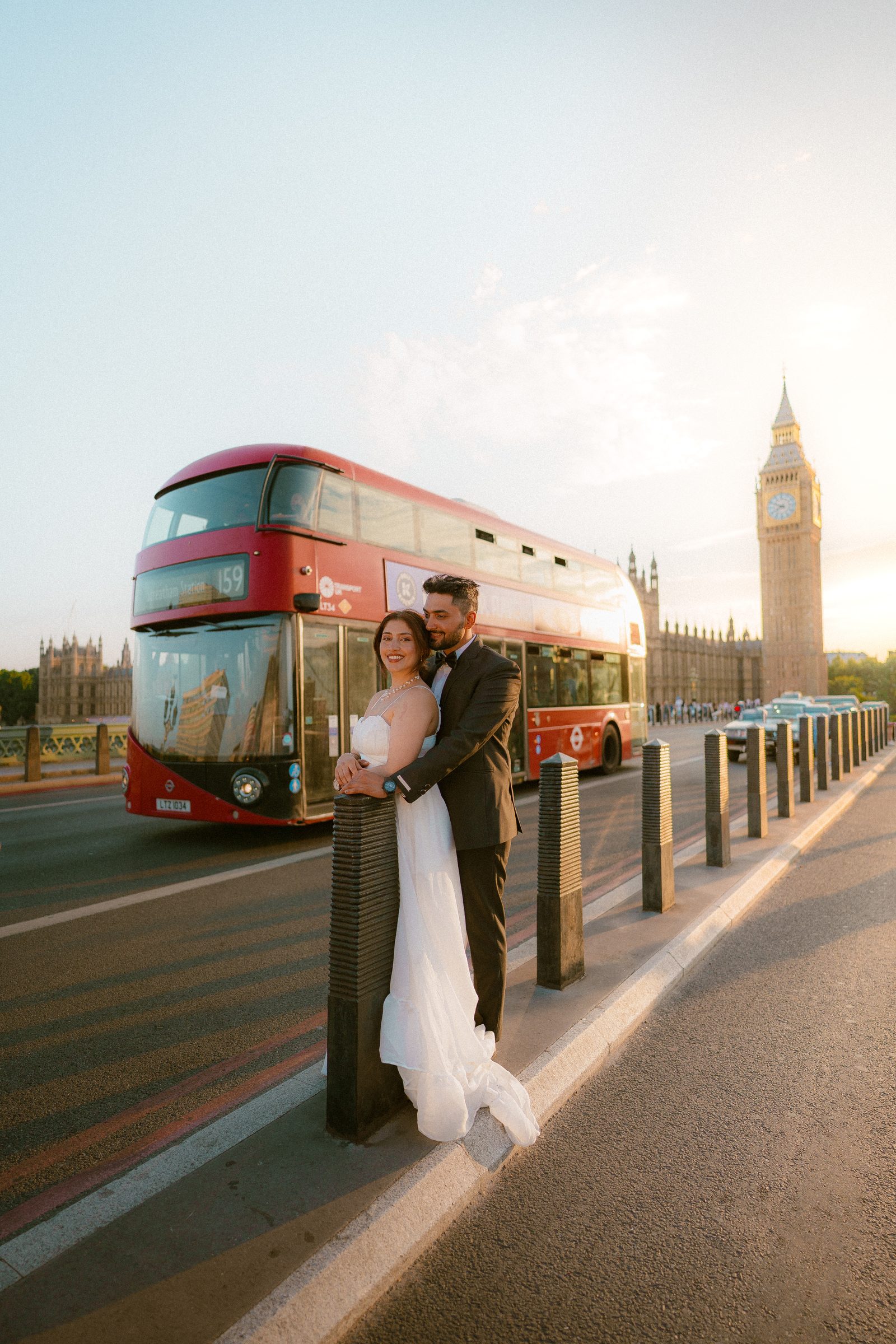 A couple in wedding clothes standing close on Westminster Bridge as a red double-decker bus passes behind them