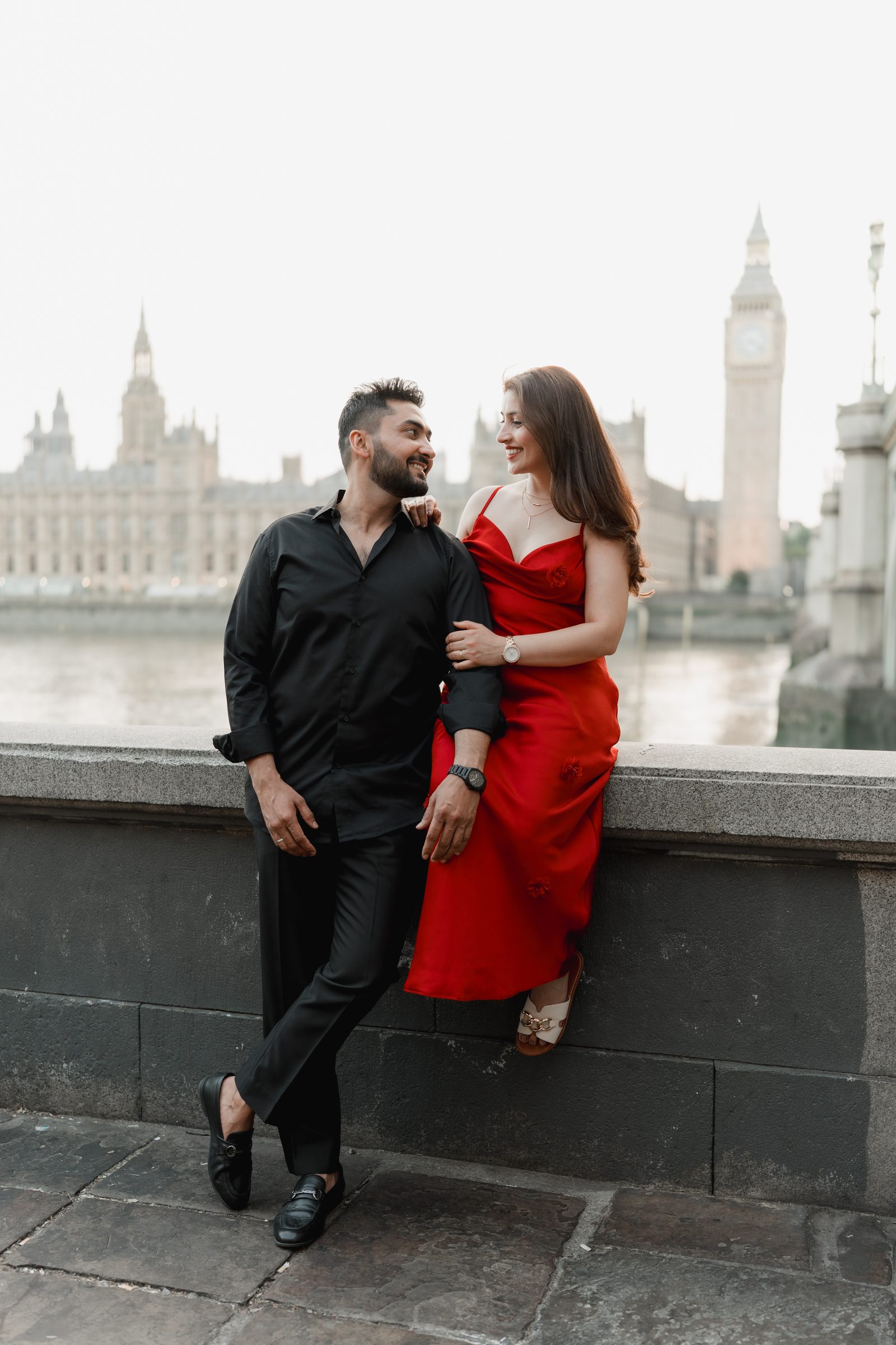 A couple sitting on the river wall laughing together, the Houses of Parliament and a pink sunset sky behind them