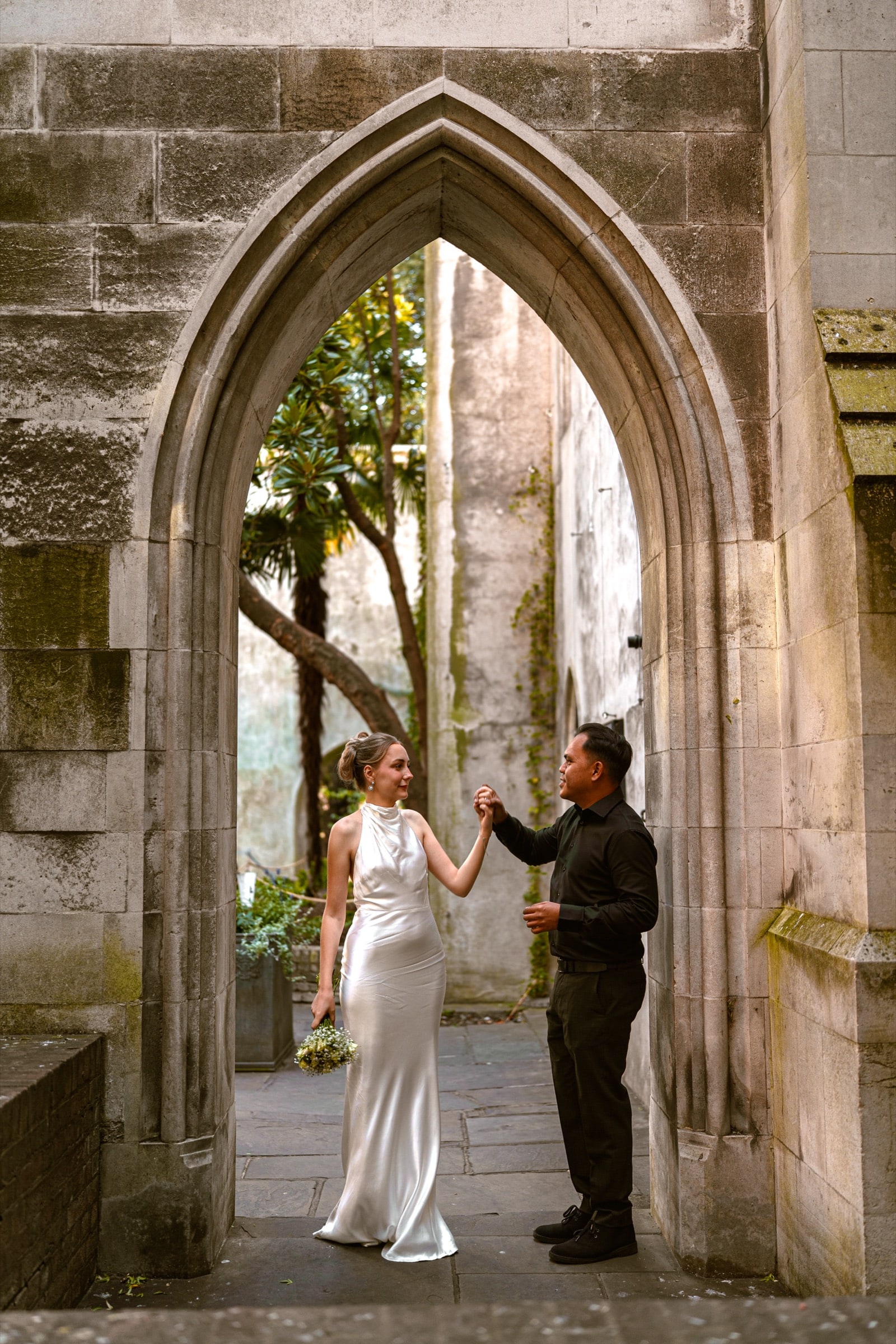 A couple holding hands beneath a gothic arch in the ruined church garden of St Dunstan in the East
