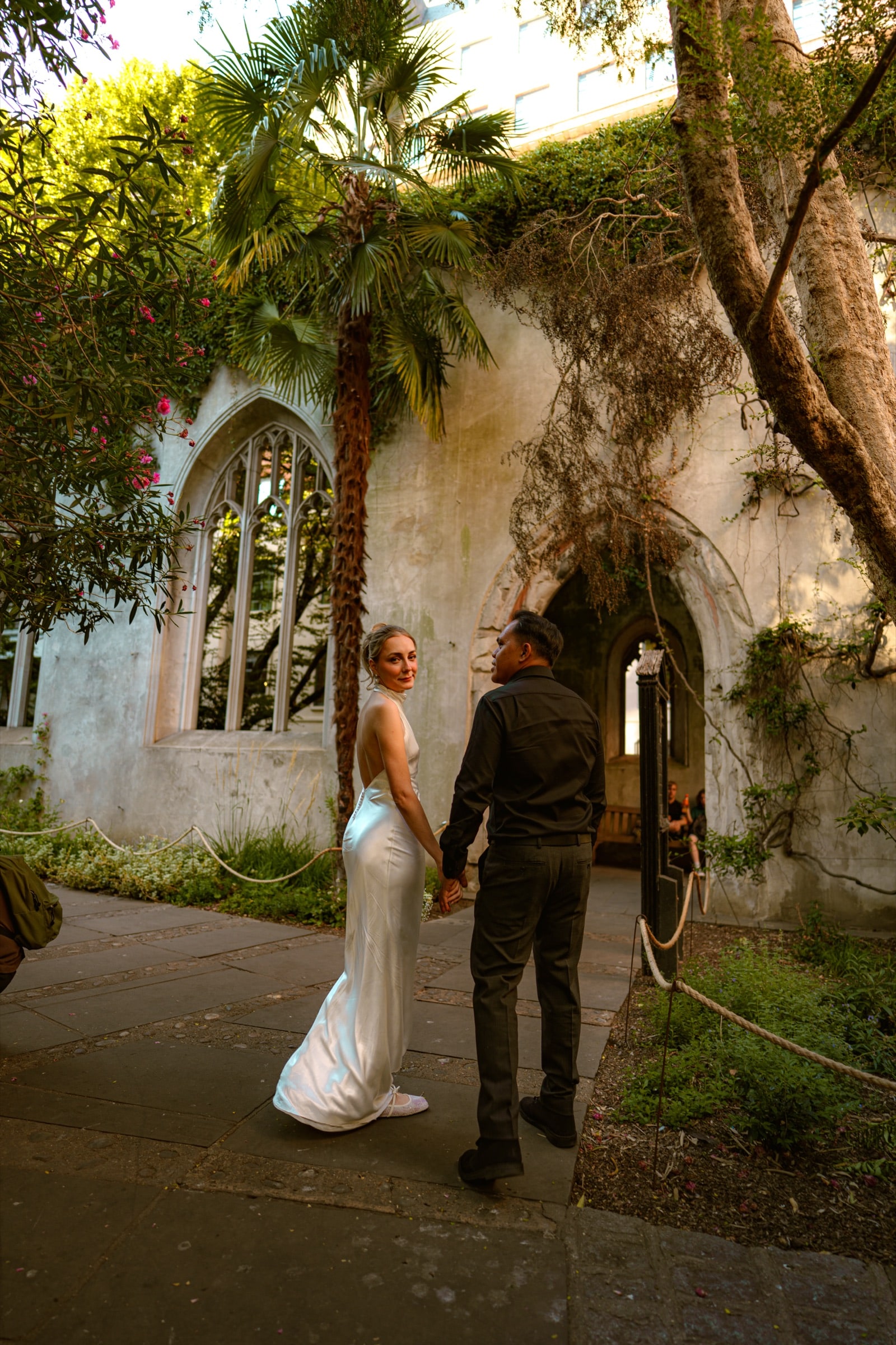 A couple walking the garden path at St Dunstan in the East, a palm tree and gothic window rising from the ruined church walls