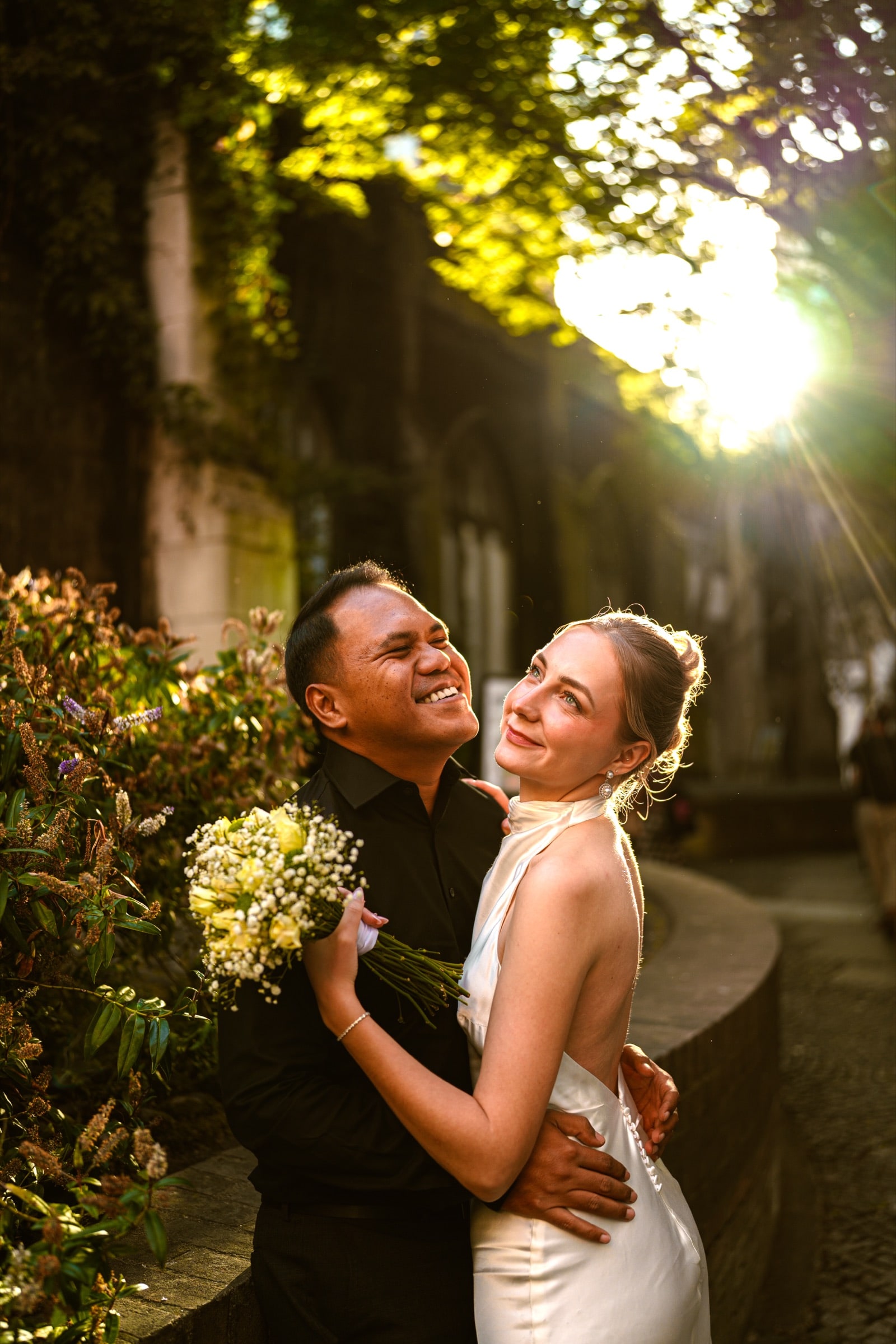 A couple laughing together at St Dunstan in the East as low golden-hour sun breaks through the trees behind them
