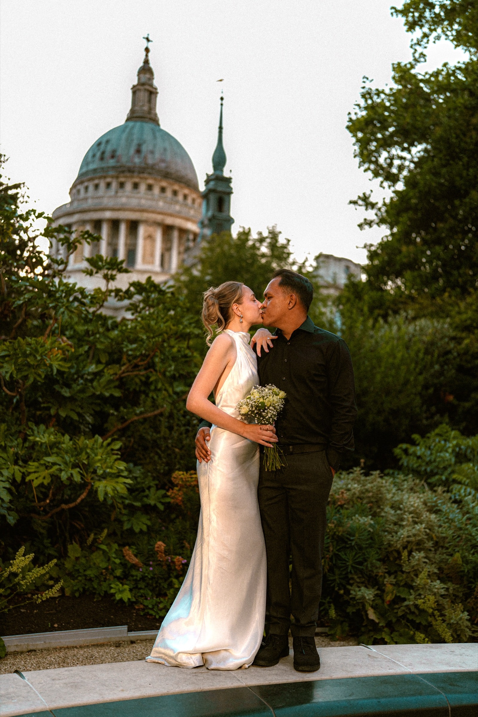 A couple kissing in the Festival Gardens at dusk, the dome of St Paul's rising behind the trees
