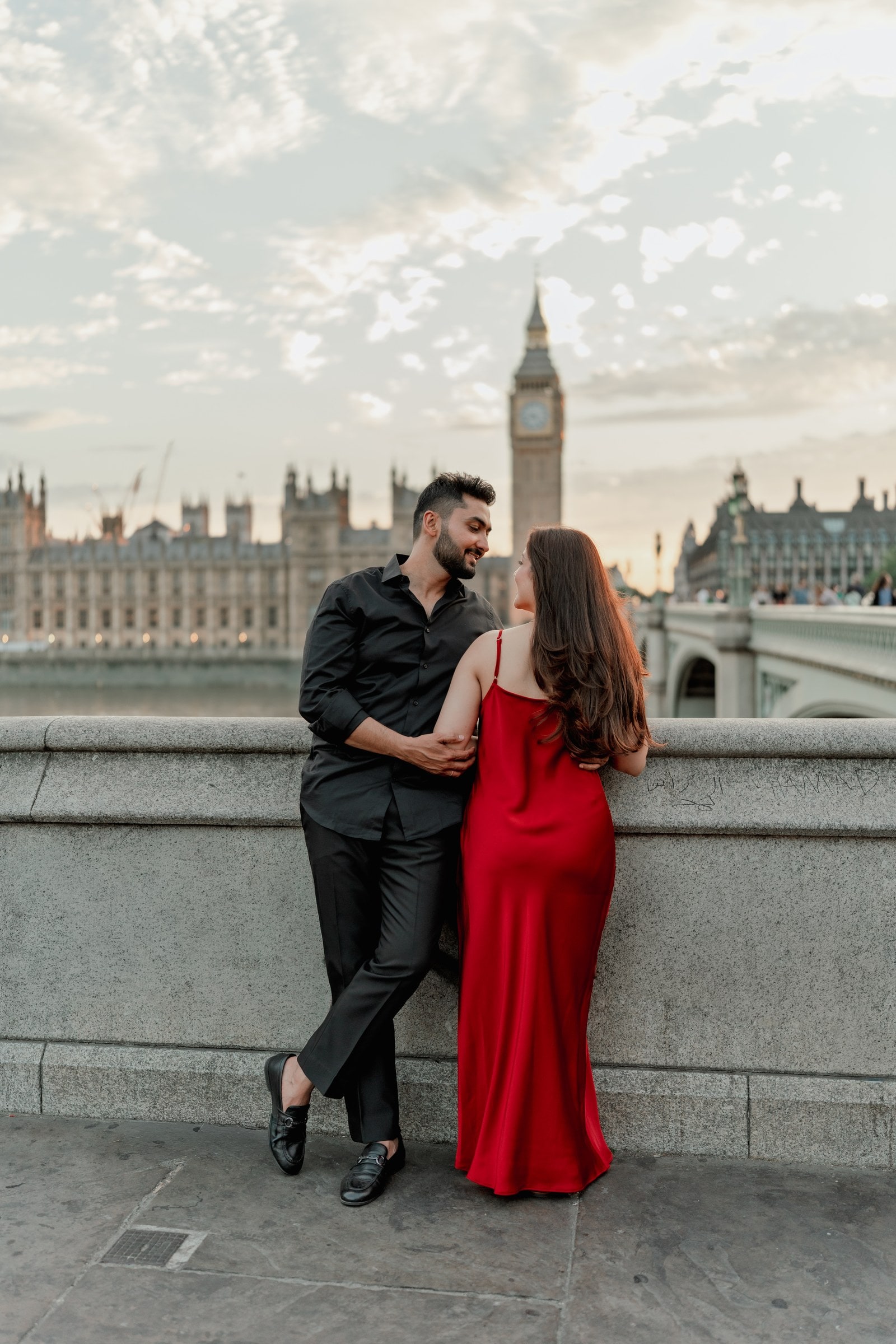 A couple leaning together against the river wall at sunset, Big Ben and an evening sky behind them