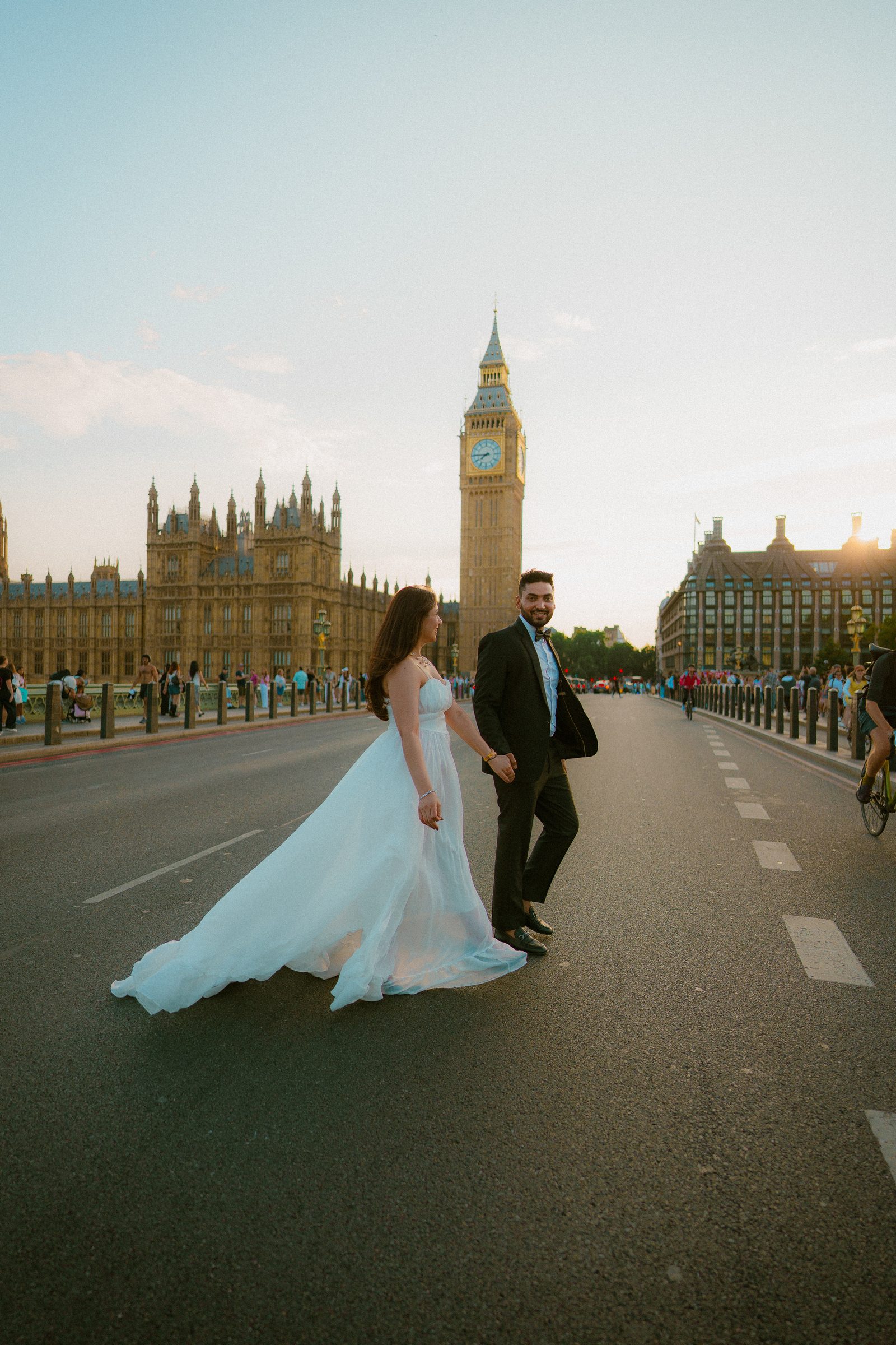 A couple crossing the road on Westminster Bridge hand in hand, her long white train sweeping the tarmac, Big Ben ahead
