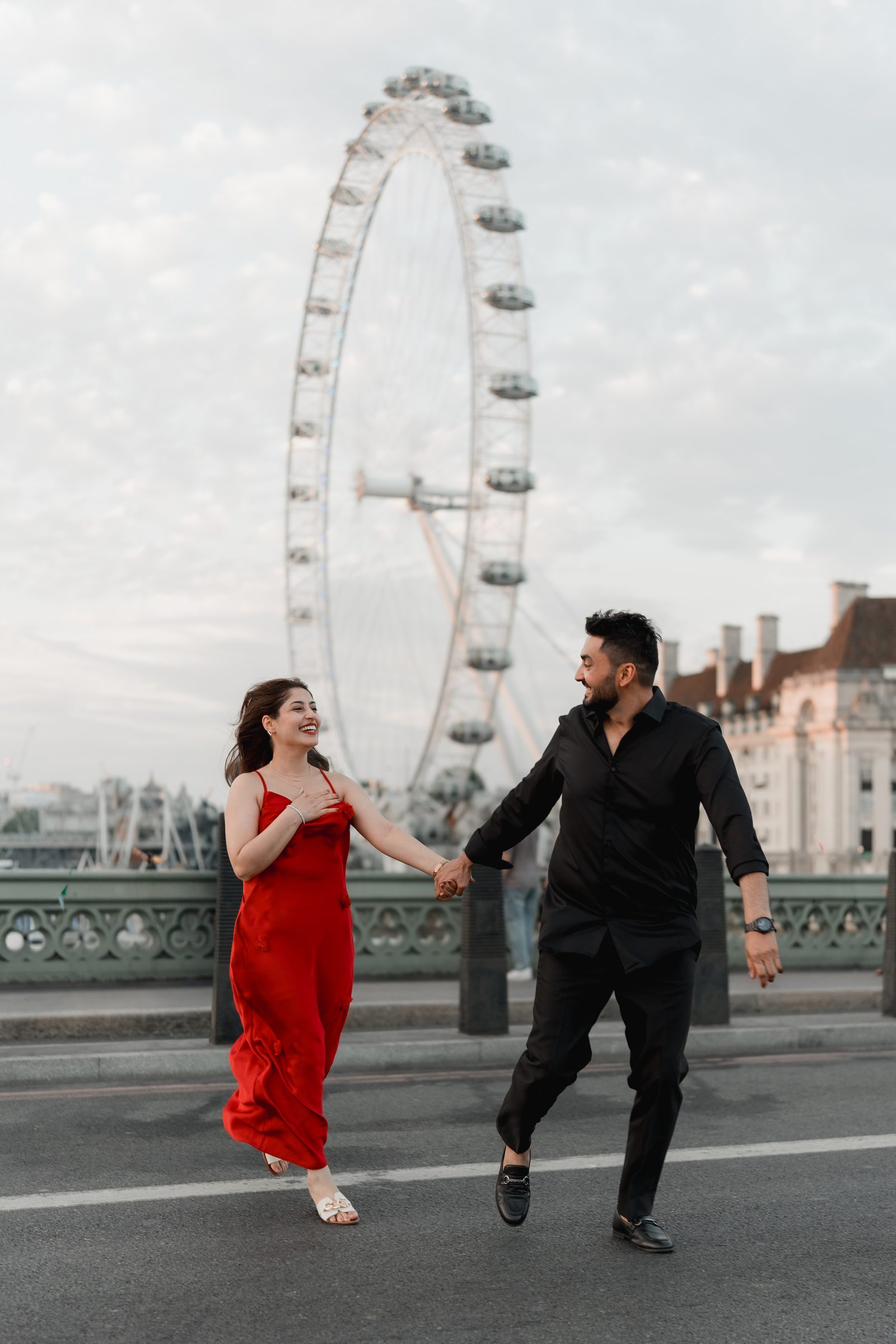 A couple walking hand in hand across Westminster Bridge in the evening, her red dress moving with the breeze