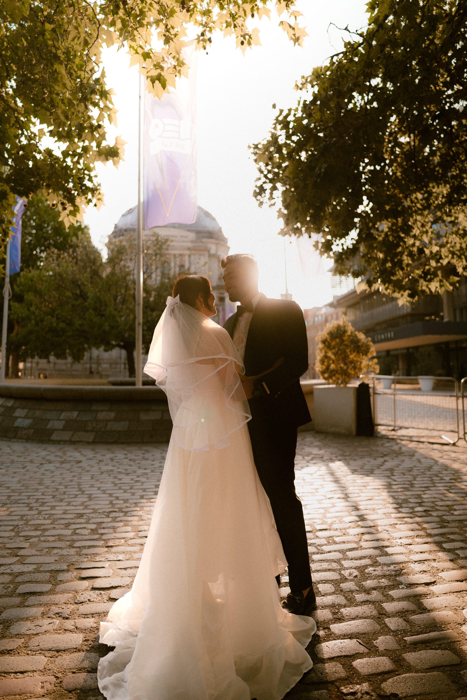 A couple in bridal whites and a black suit standing on backlit cobblestones, her veil glowing in the low evening sun in Westminster