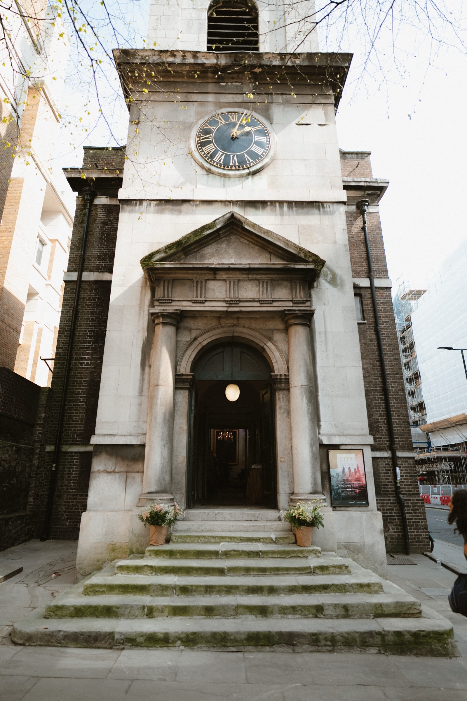 The Georgian stone facade, pedimented portico and clock tower of All Hallows-on-the-Wall, with City of London glass towers beside it