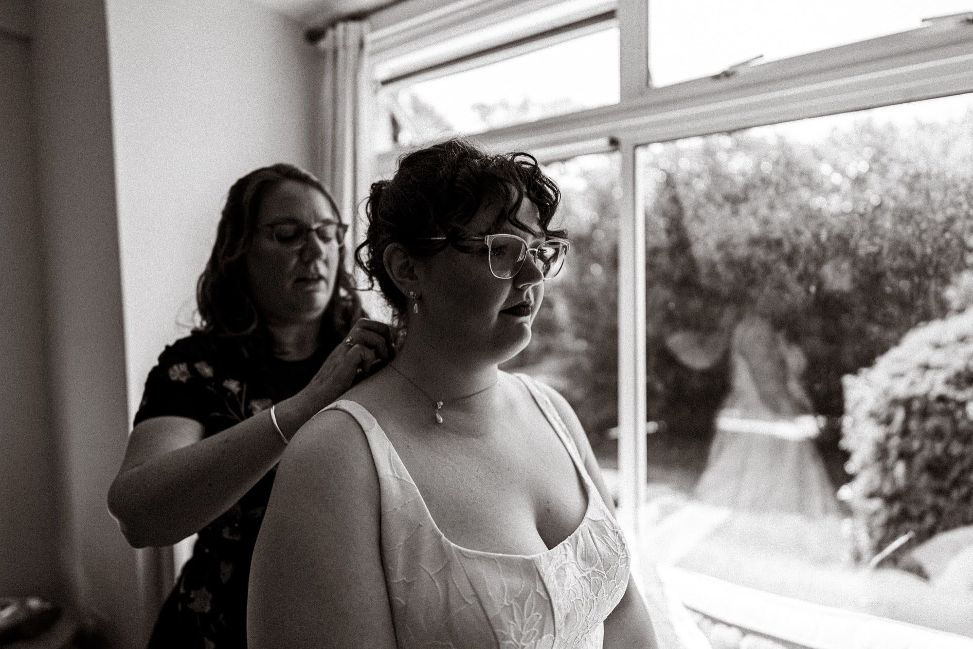 Black and white photograph of the bride having her dress fastened by the window, her gown reflected in the glass, before the All Souls, Langham Place wedding