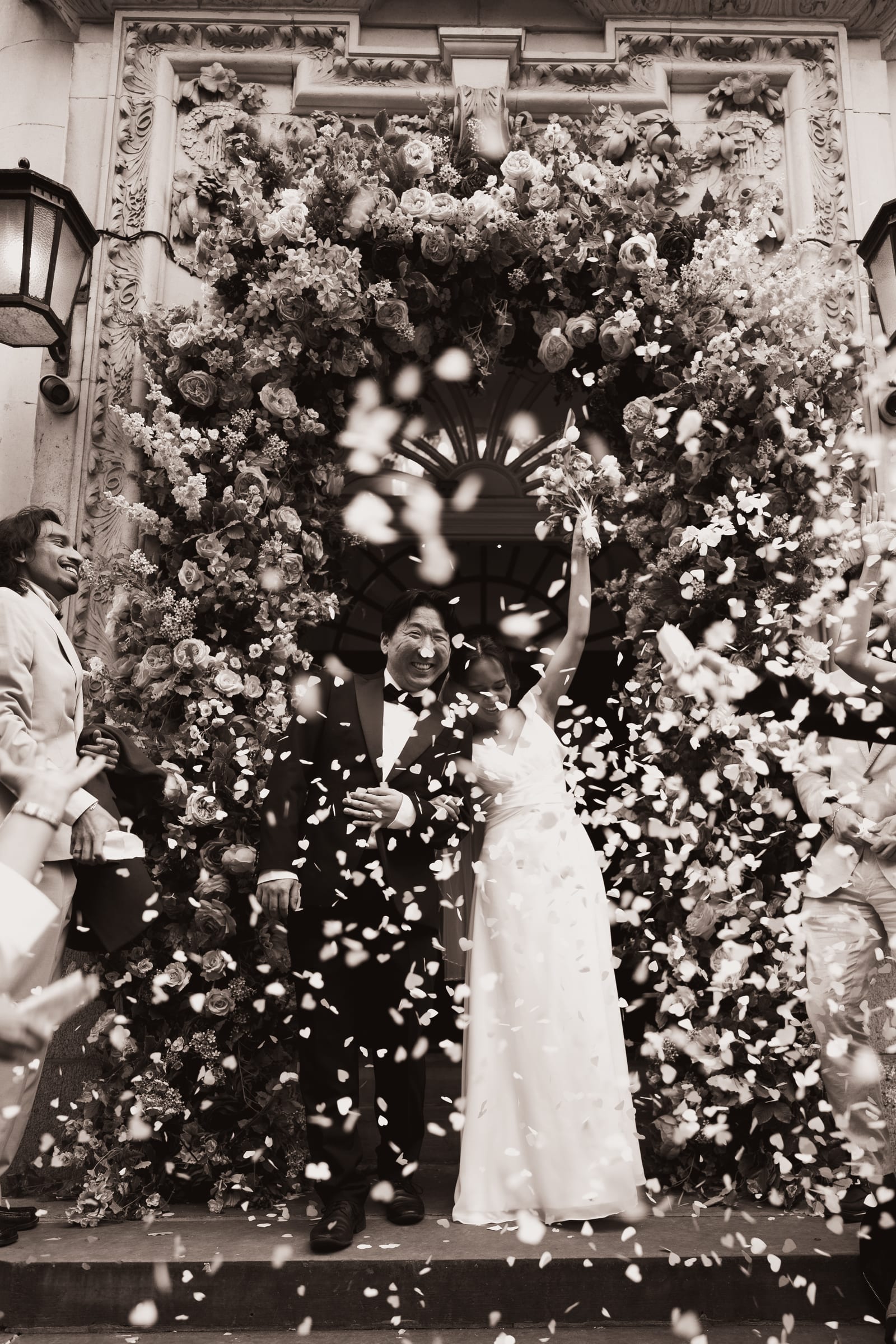 Newlyweds throwing confetti at the top of the steps beneath the floral arch at Chelsea Old Town Hall