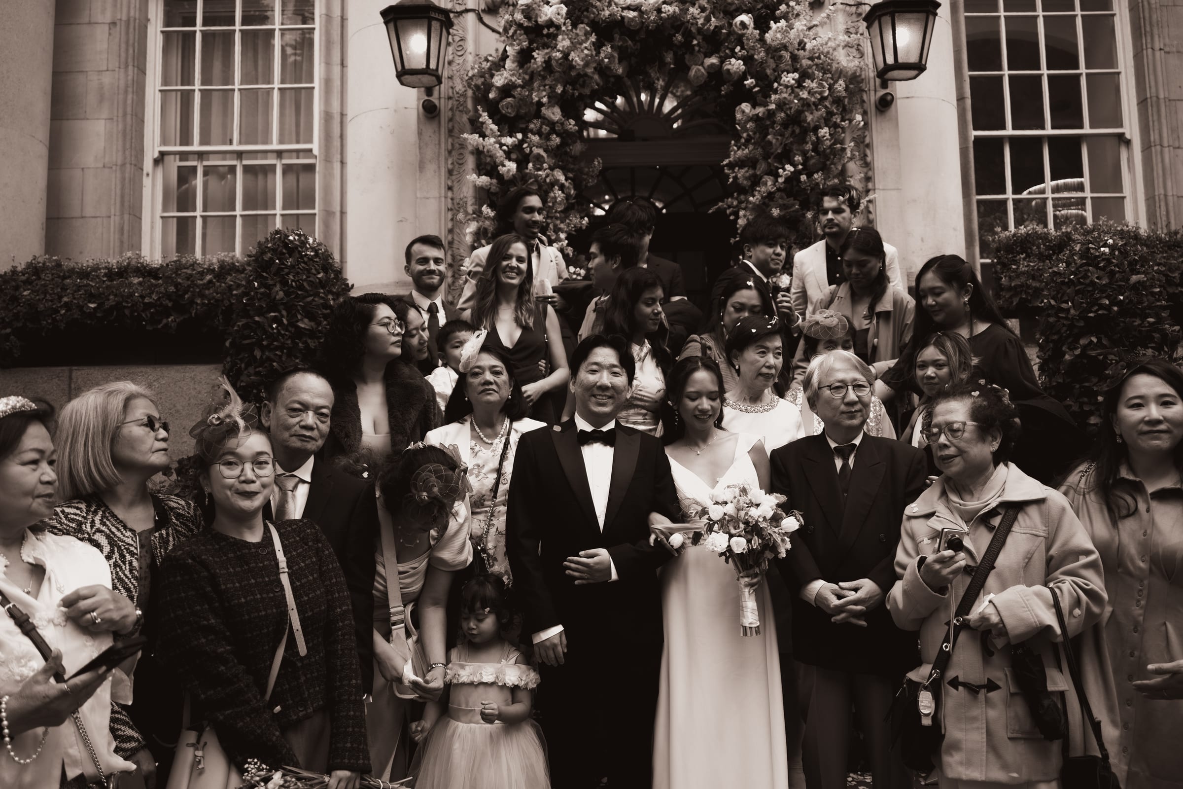 Sepia group photograph of the wedding party on the front steps of Chelsea Old Town Hall beneath the floral arch