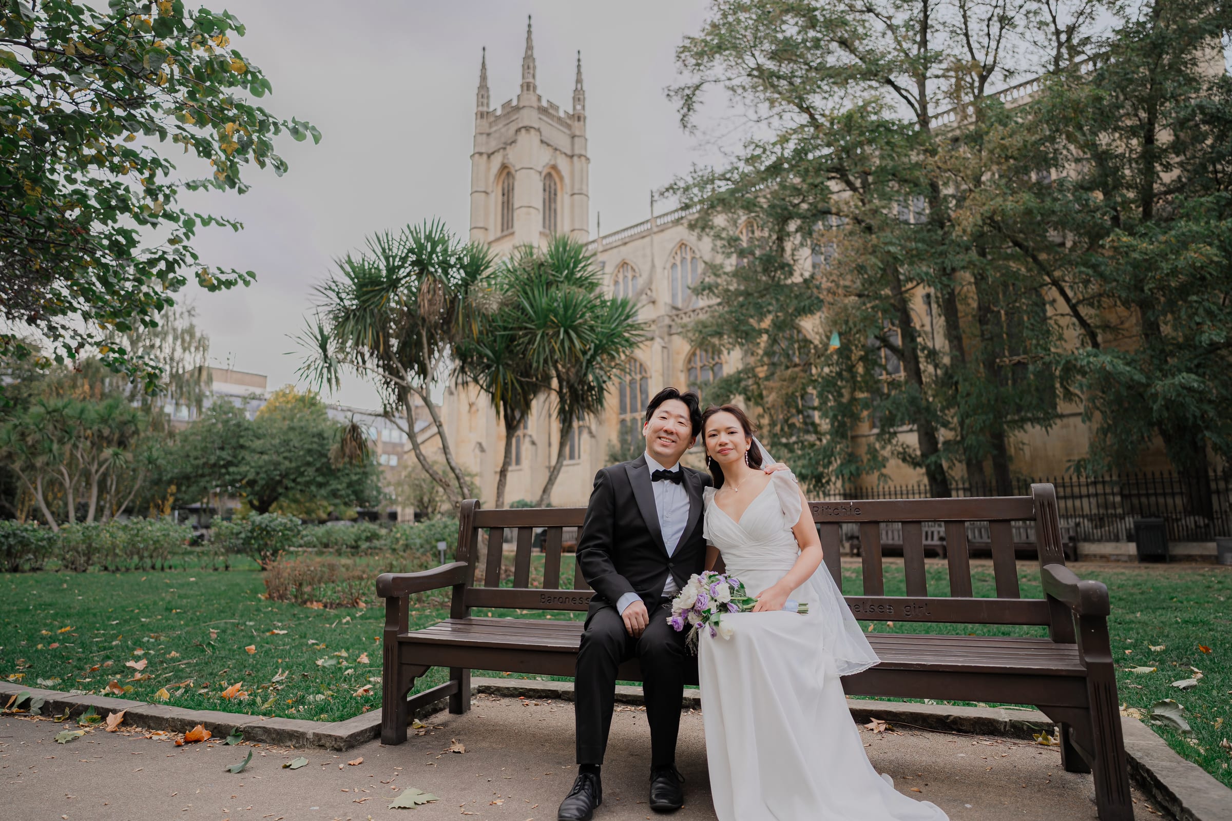 Bride and groom on a bench in St Luke’s Gardens, Chelsea, the Gothic church rising behind them