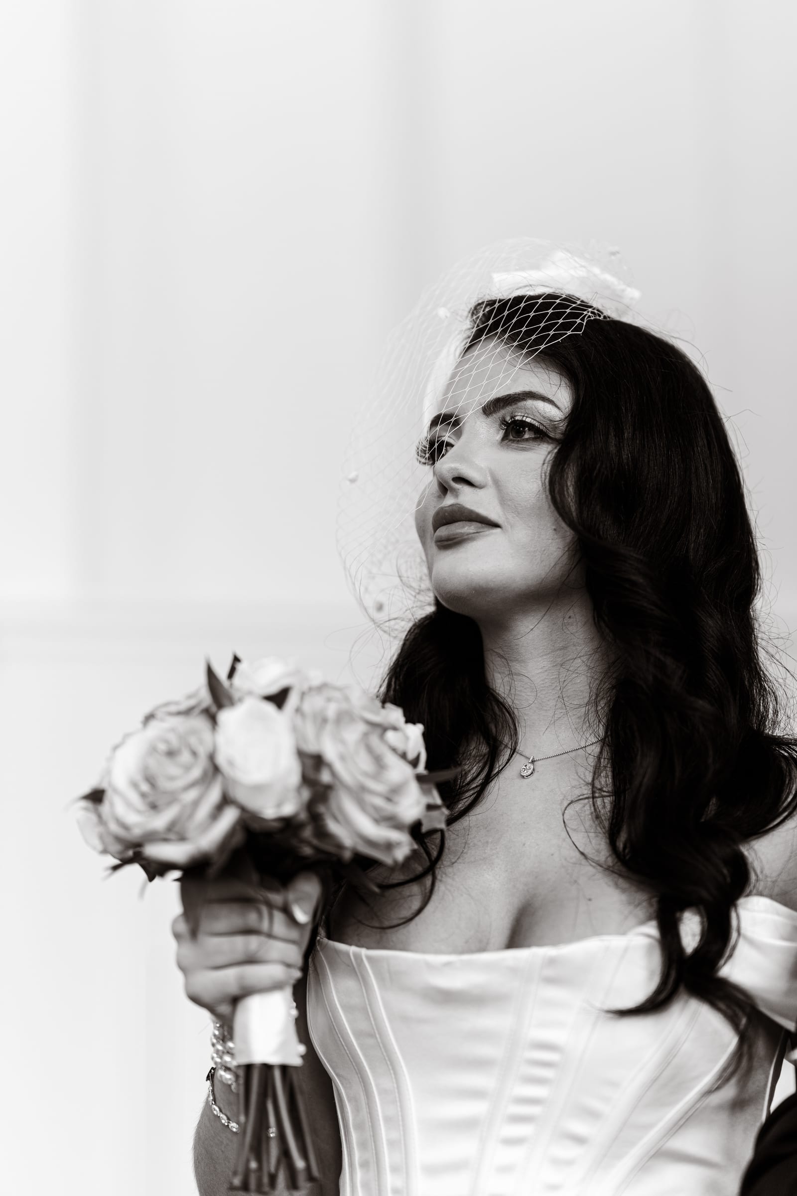 Black and white portrait of a bride in a corseted gown and birdcage veil holding her rose bouquet at Cheshunt Register Office