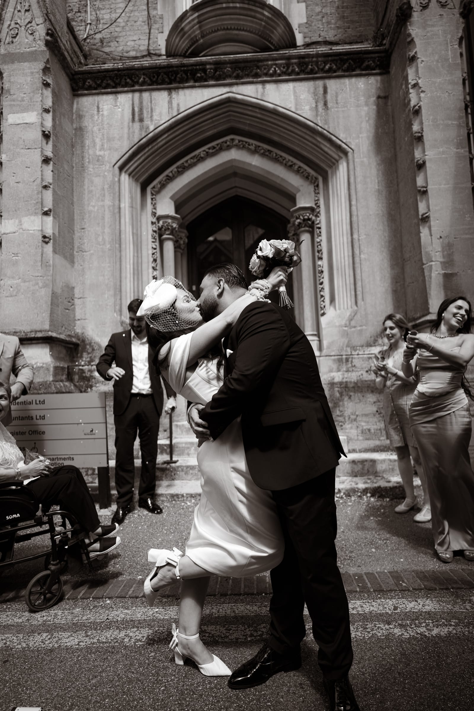 A groom lifting his bride into a kiss as guests throw confetti outside Cheshunt Register Office, in black and white