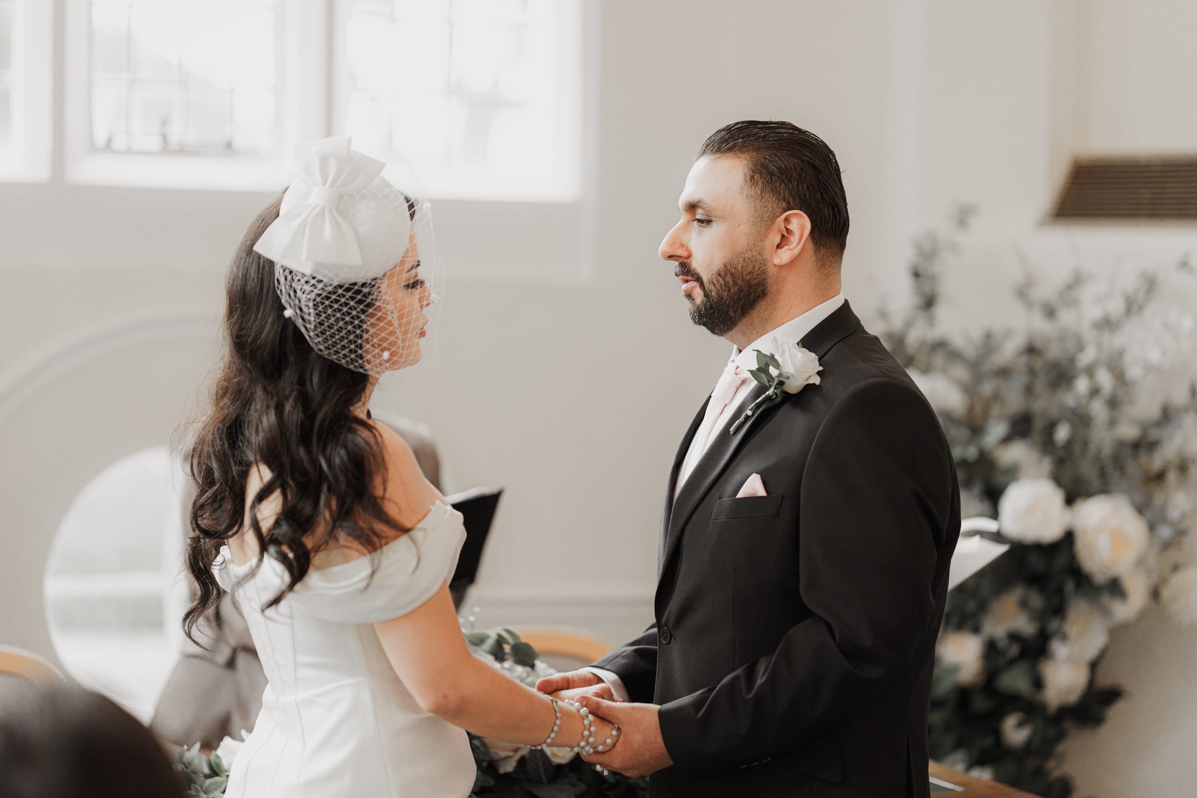 A couple holding hands during their vows in the Octagonal Suite at Cheshunt Register Office, white flowers behind them