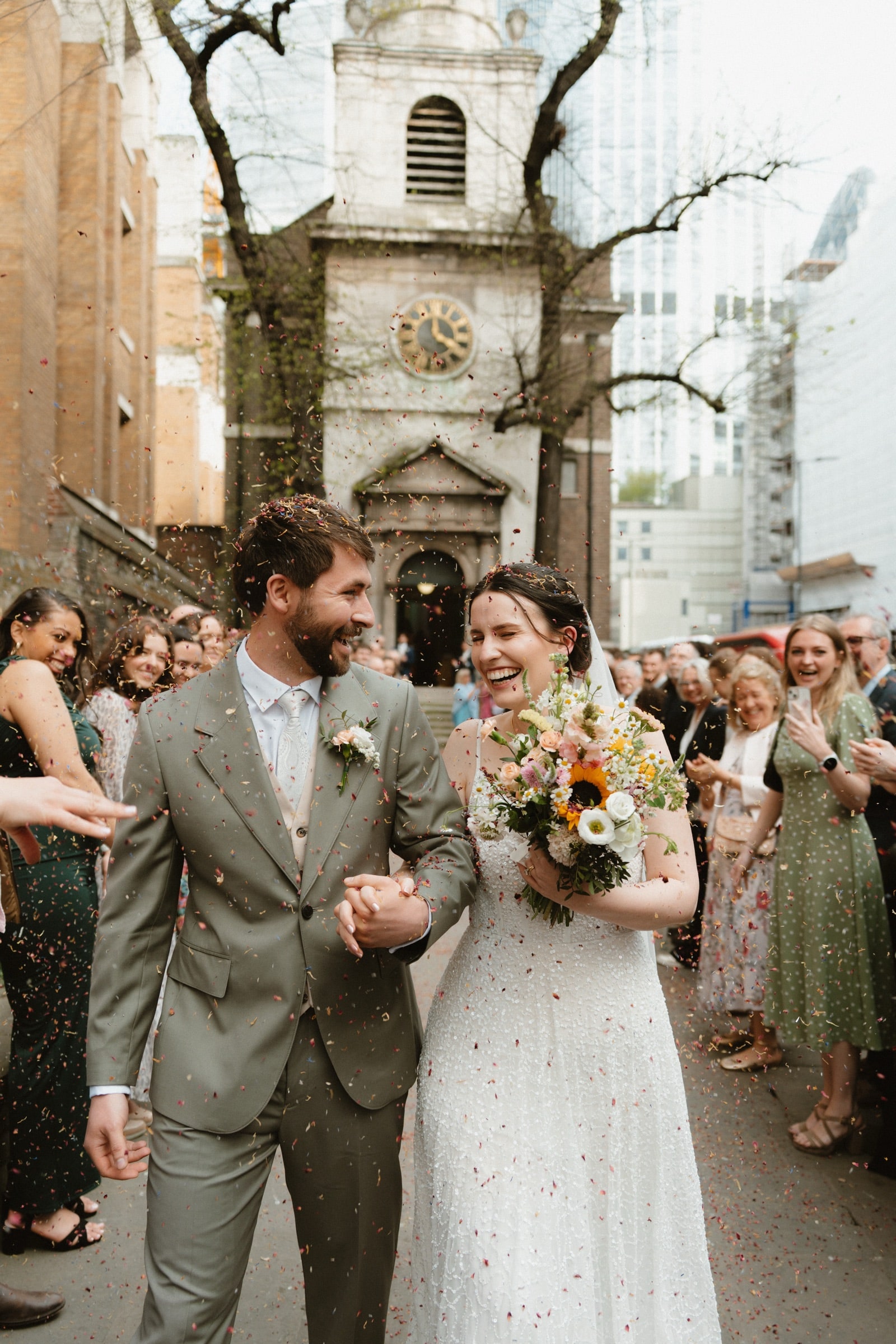 Couple laughing through a confetti exit outside All Hallows-on-the-Wall, a City of London glass tower rising behind