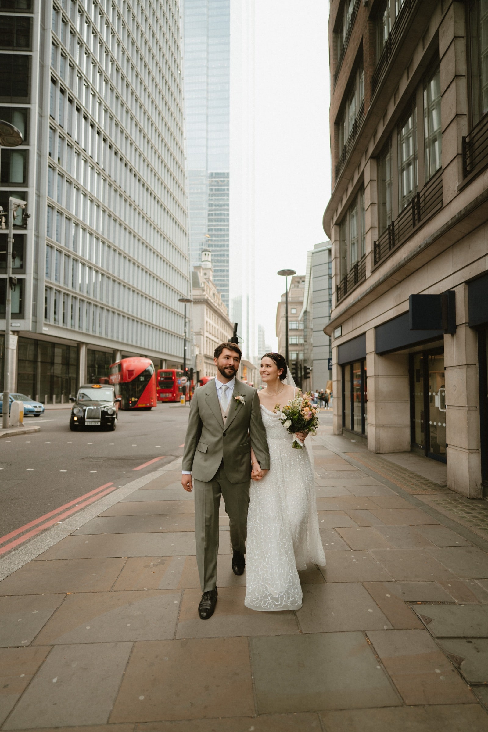 Bride and groom walking a City of London street between tall buildings, a red bus and black cab behind