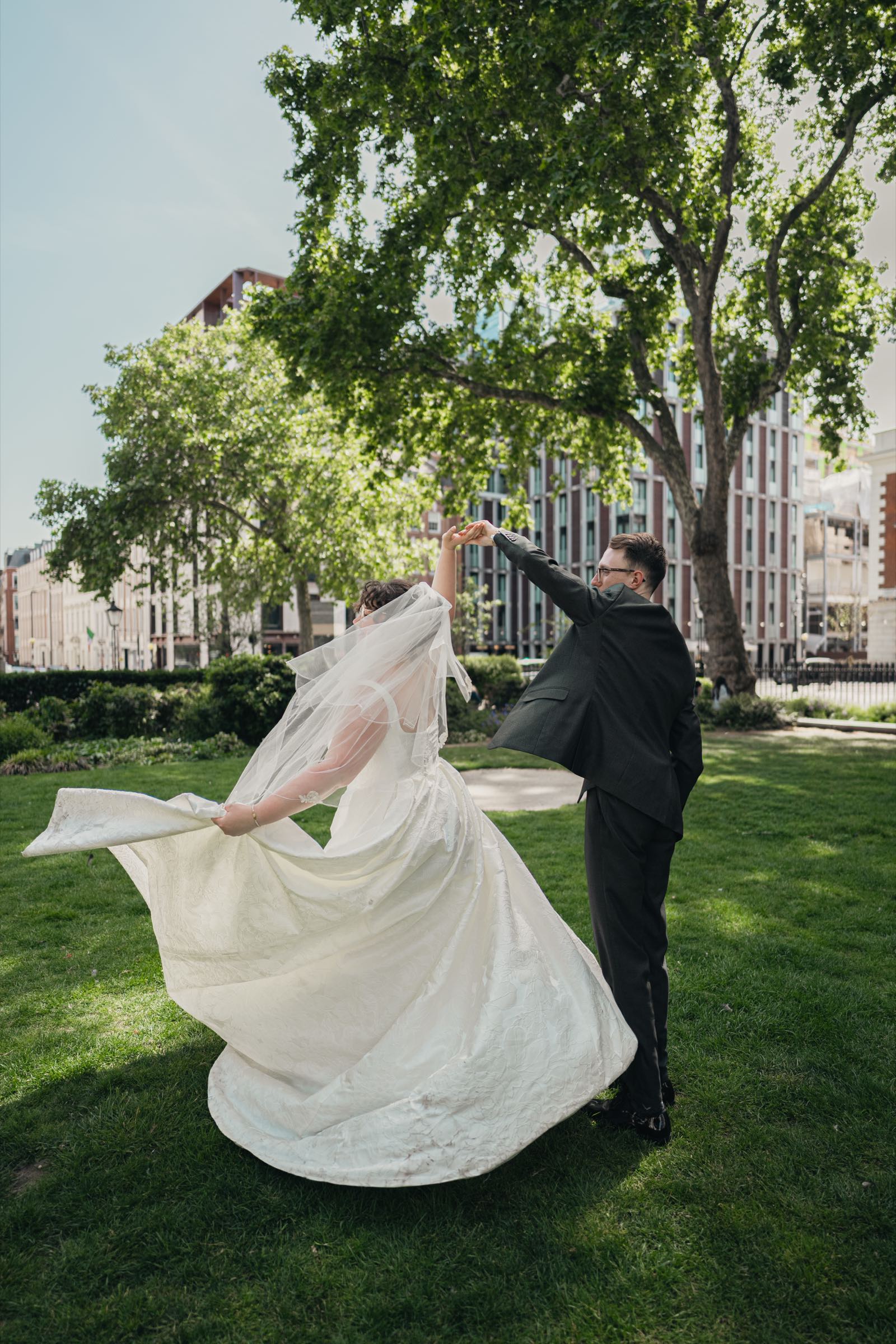 A groom twirling his bride, her dress and veil flaring out, in a London park after their ceremony