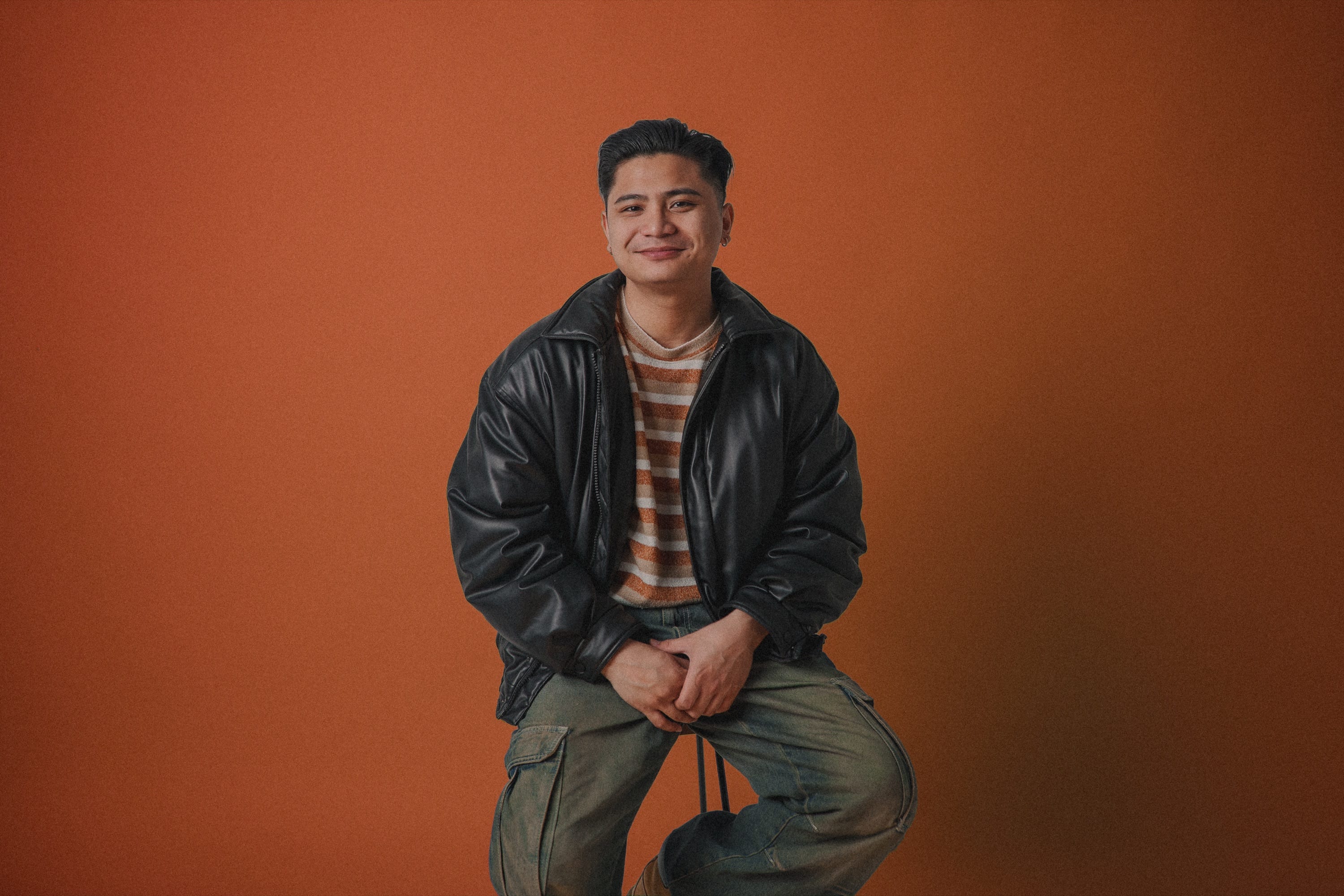 John Roberto, sitting on a stool and smiling against an orange backdrop
