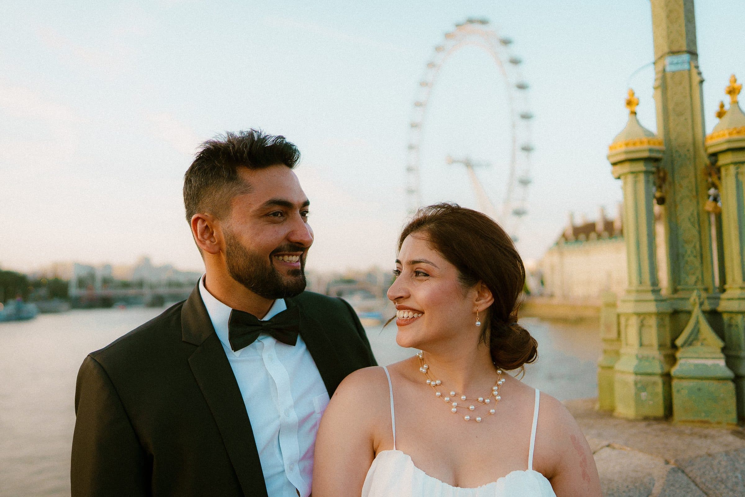 A couple in evening wear standing together beneath an ornate lamp post on Westminster Bridge with the London Eye behind