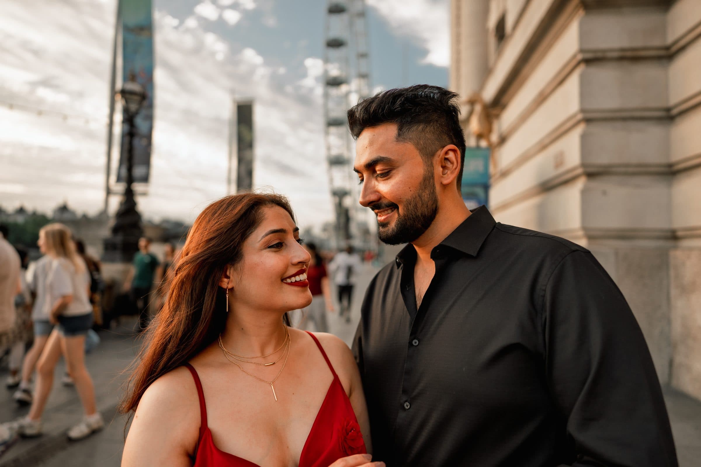 A couple laughing close together on the South Bank at golden hour, she in a red dress, during a London engagement shoot