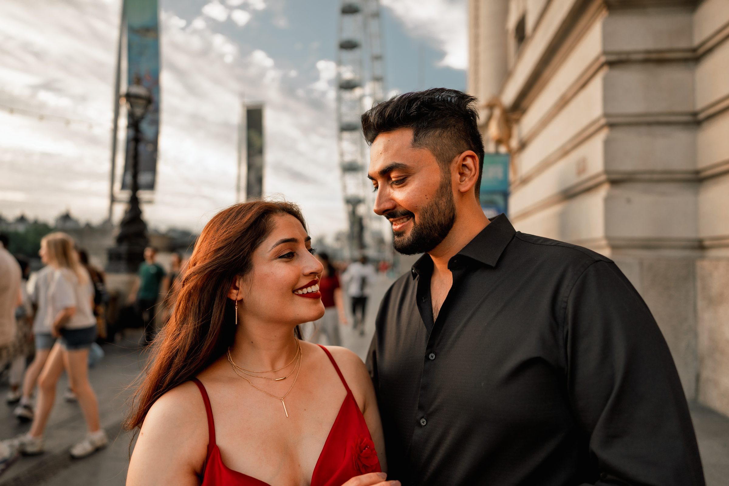 A couple laughing close together on the South Bank at golden hour, she in a red dress, during a London engagement shoot